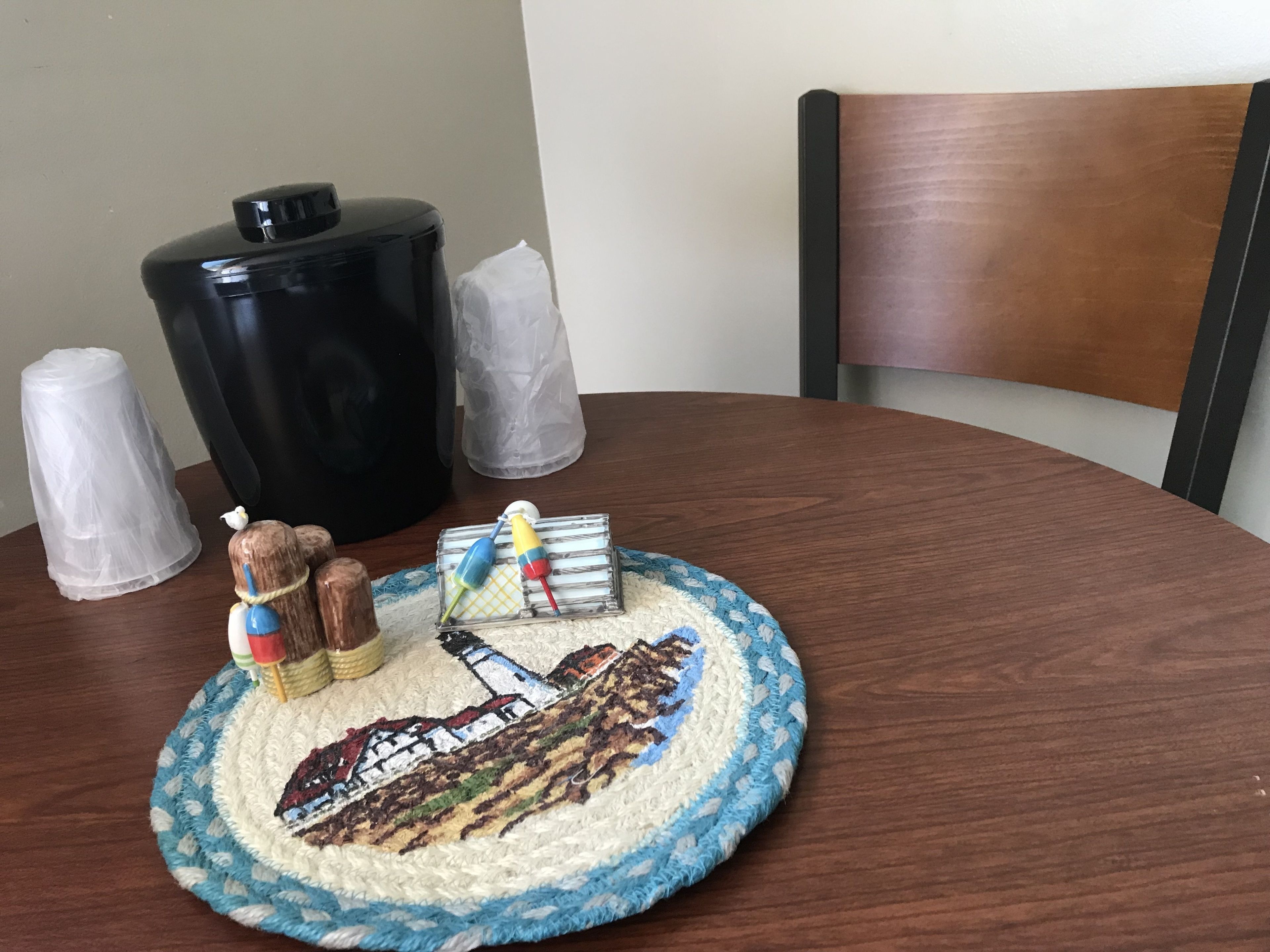 A round wooden table with a crocheted placemat featuring a landscape design, a black container, and two clear plastic candles.