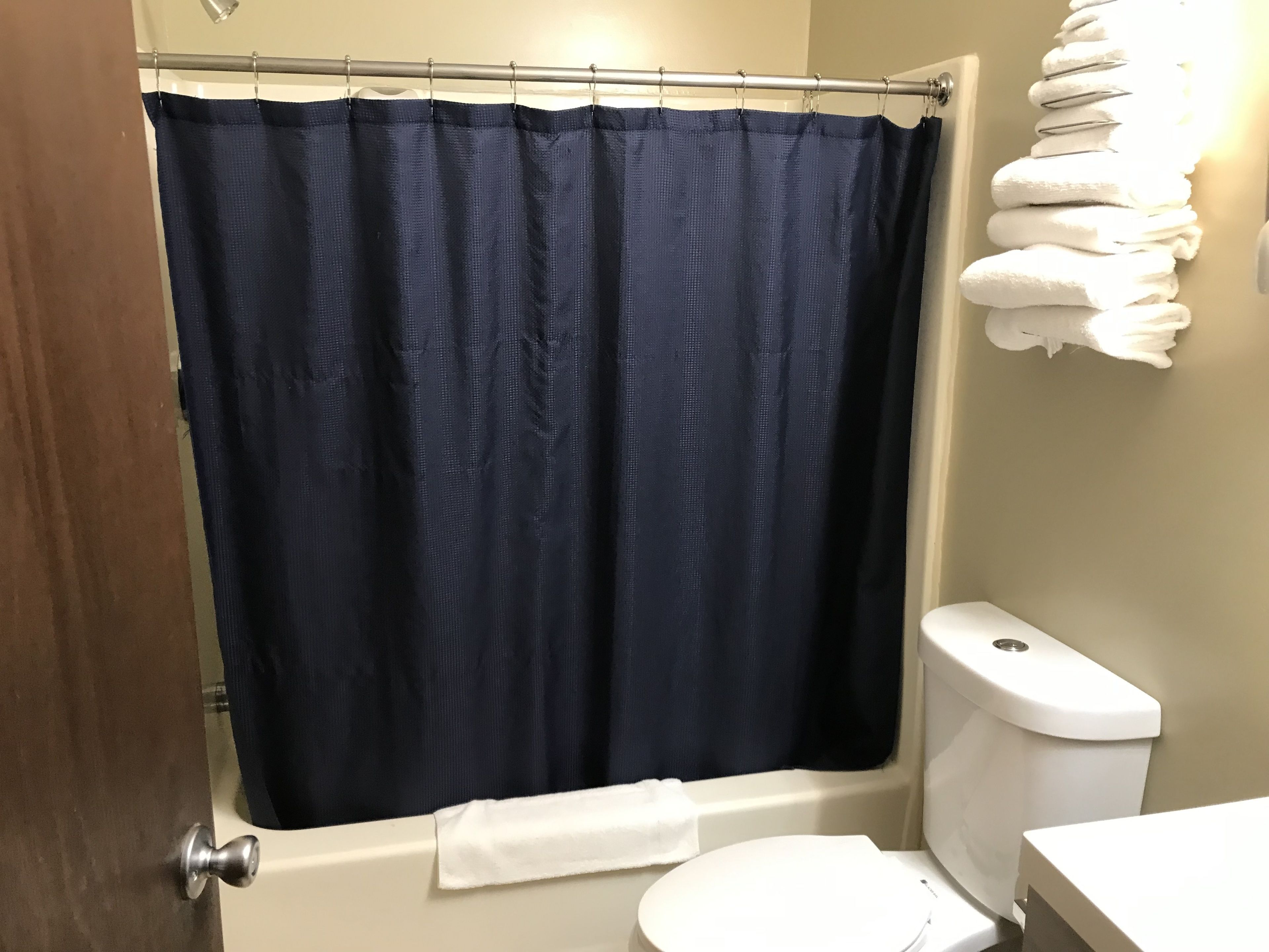 Un baño con cortina de ducha azul oscuro, inodoro blanco y una pila de toallas blancas en la pared.