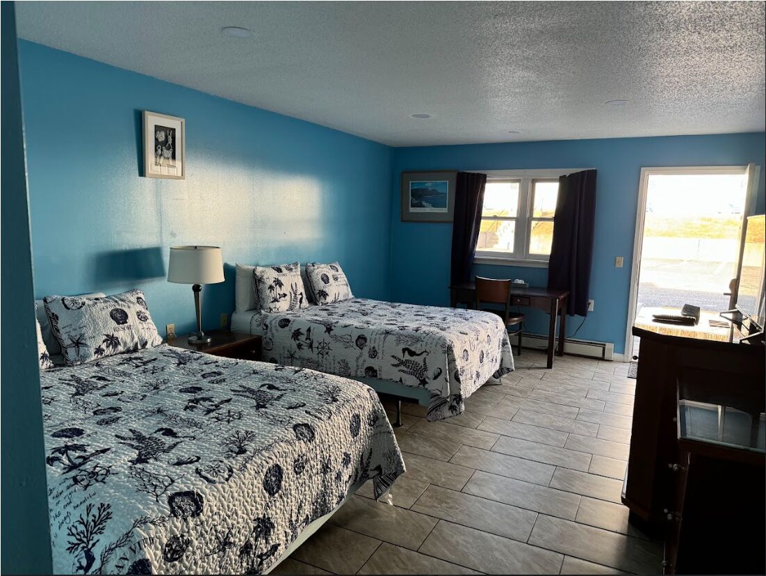Quarto com duas camas, uma escrivaninha, uma cadeira, um abajur e uma janela com cortinas.
