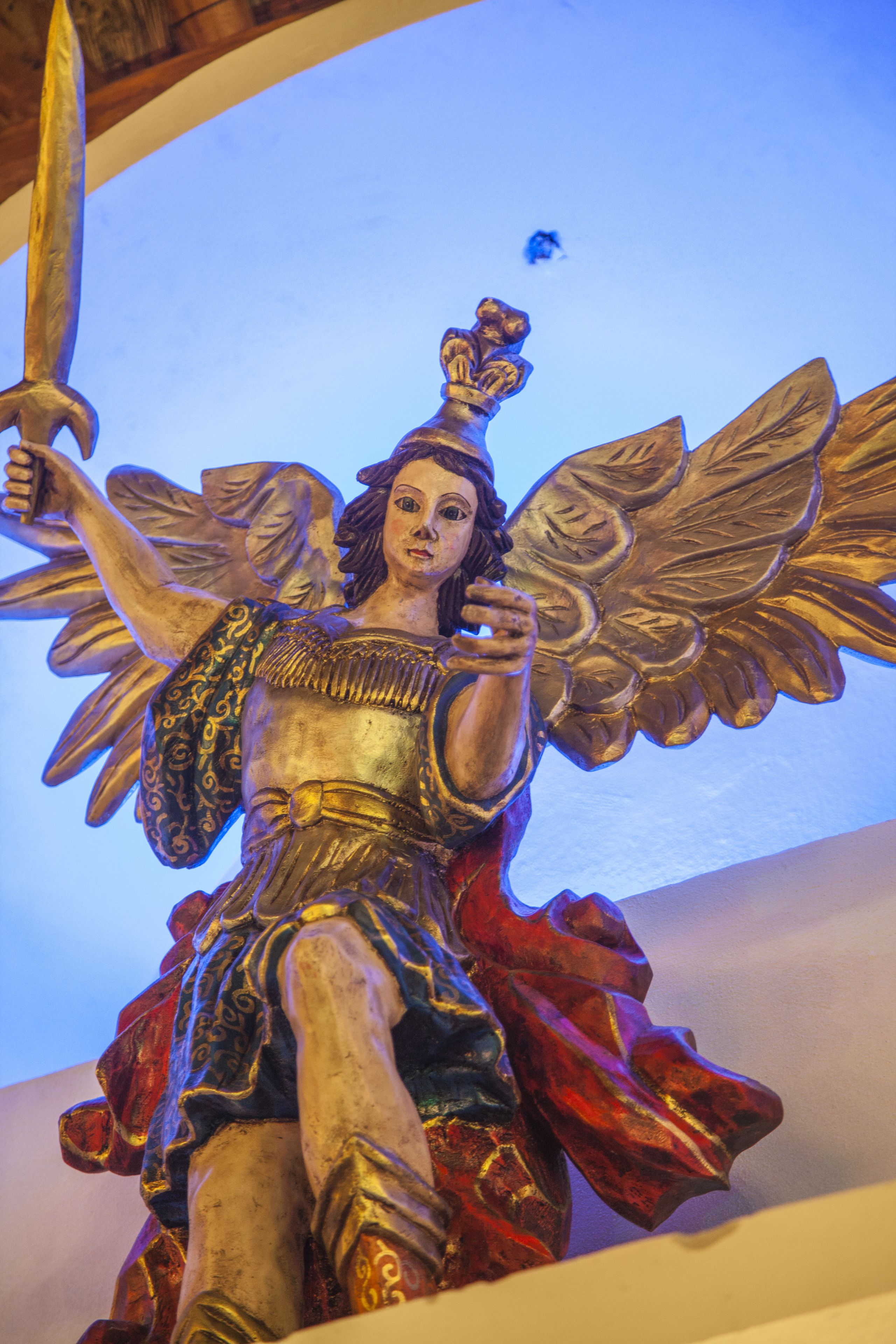 A statue of an angel with wings and a sword, positioned in a room with a curved ceiling and a blue light in the background.