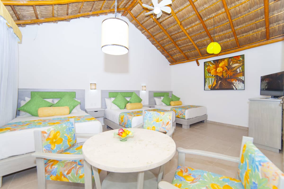A hotel room with a thatched roof, multiple beds with green and yellow pillows, a round dining table, and a TV on a cabinet.