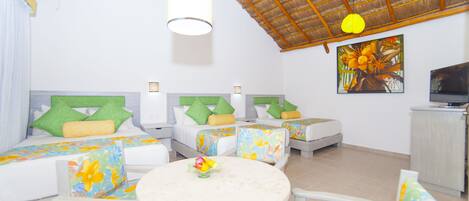 A hotel room with a thatched roof, multiple beds with green and yellow pillows, a round dining table, and a TV on a cabinet.