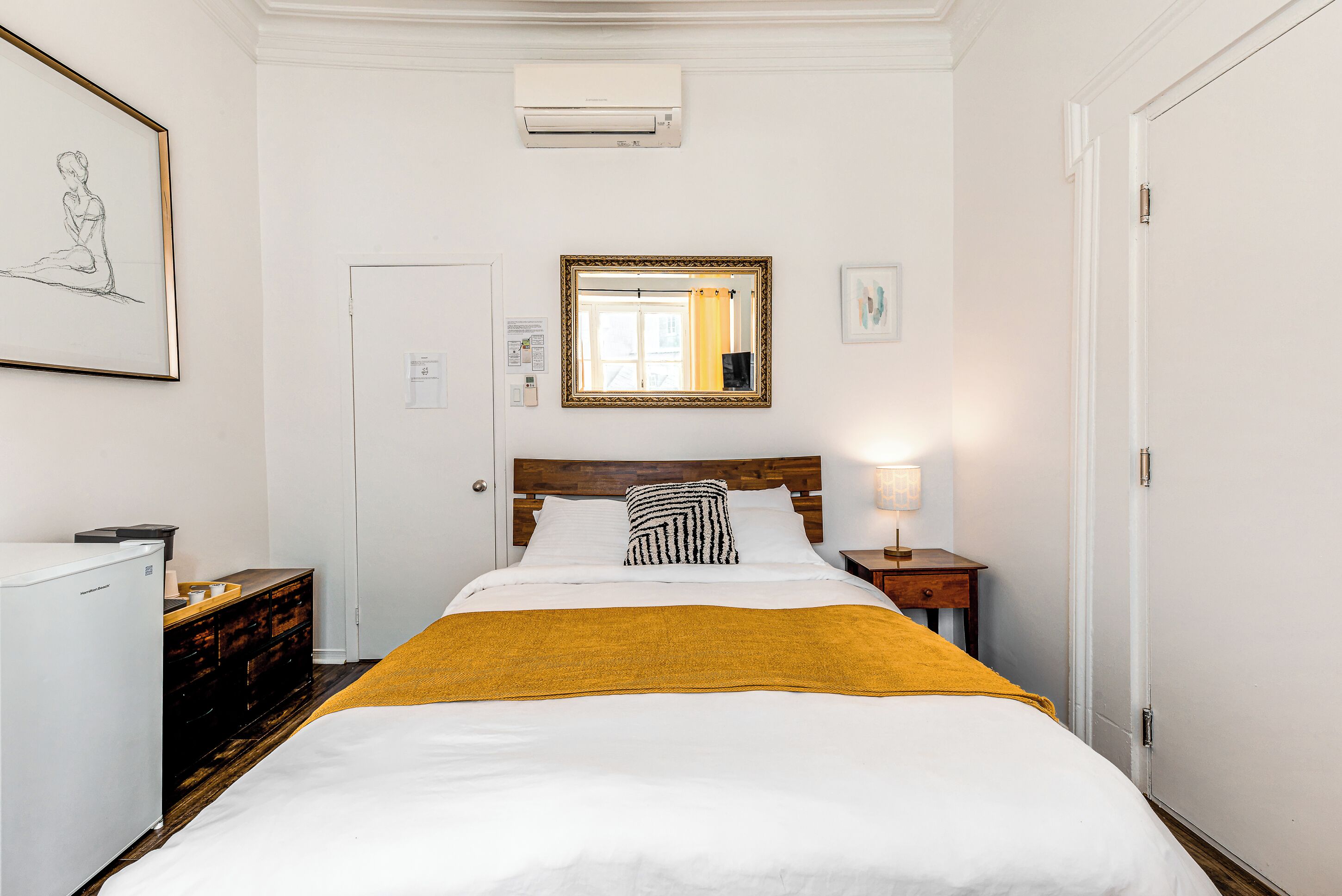 Een keurig opgeruimde hotelkamer met een bed, een nachtkastje met lamp, een dressoir en een wandairconditioner.