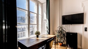 A room with a wooden desk, a black chair, a potted plant, a black refrigerator, and a TV mounted on the wall. - North House - Manoir des Remparts / Hotel Automatisé (Québec City)