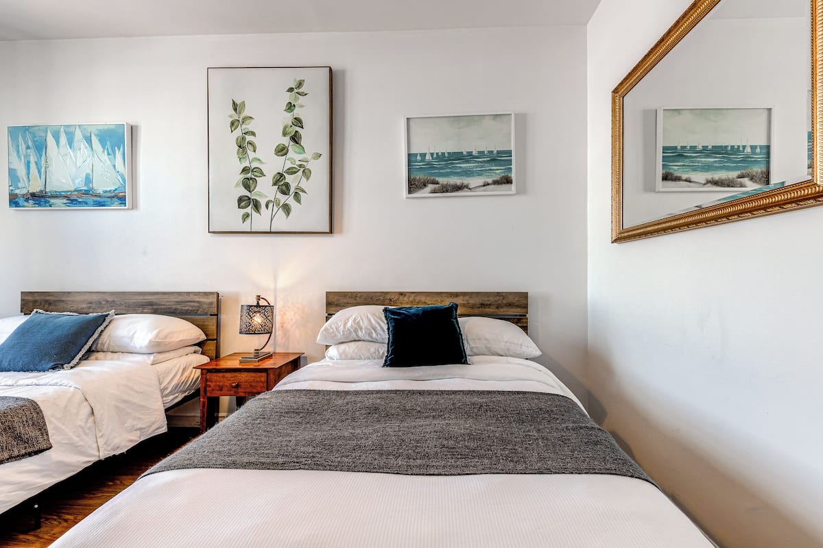 A hotel room with two beds, a nightstand, and wall art depicting sailboats and greenery.