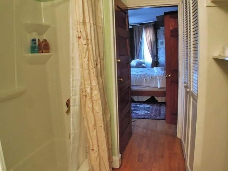 A hallway leading to a bedroom with a wooden floor, a shower area with a curtain, and a small shelf with toiletries.