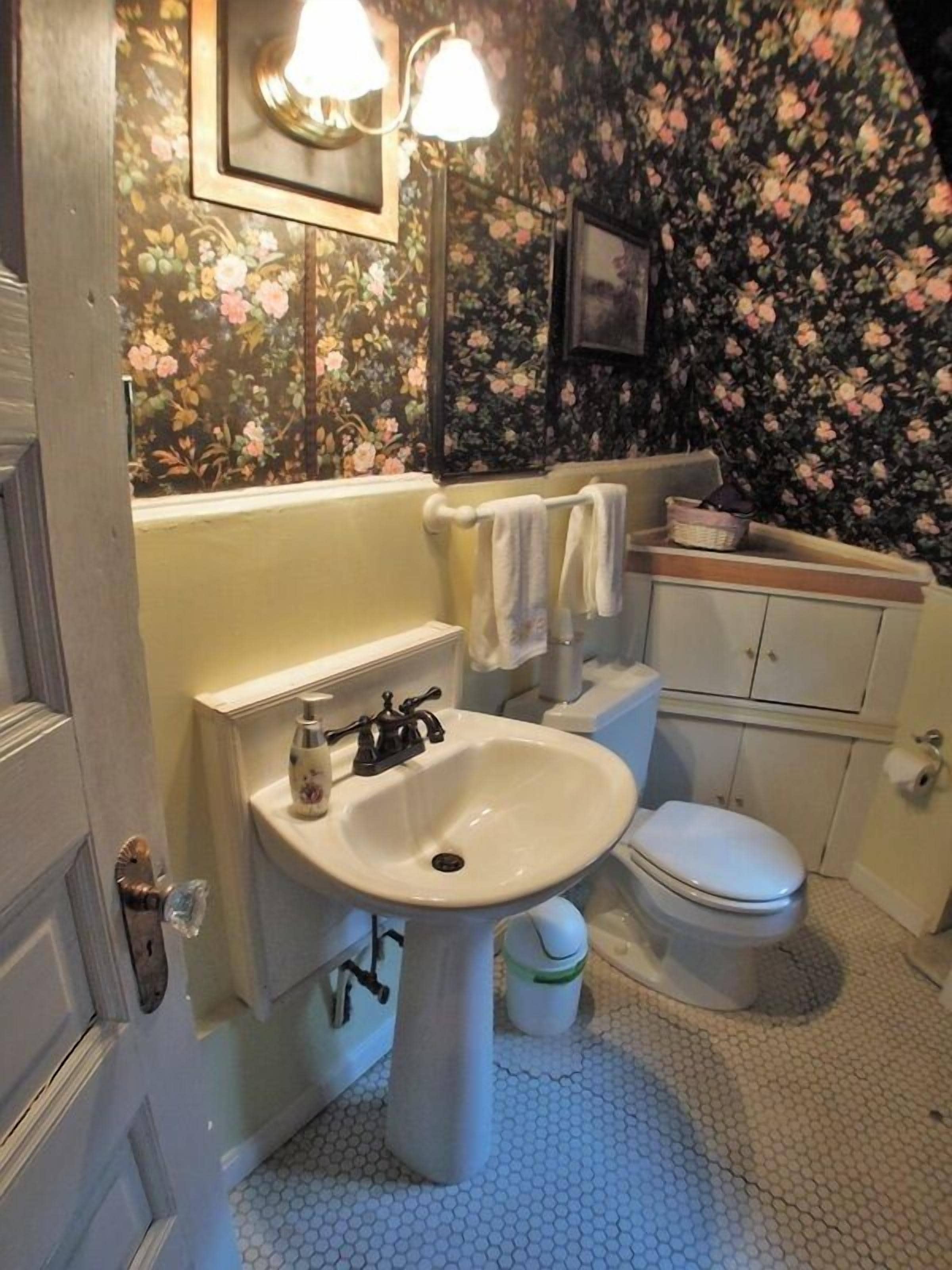 Ein Badezimmer mit einem weißen Waschbecken, einer Toilette und einem Waschtisch. Die Wände sind mit Blumenpapier tapeziert.