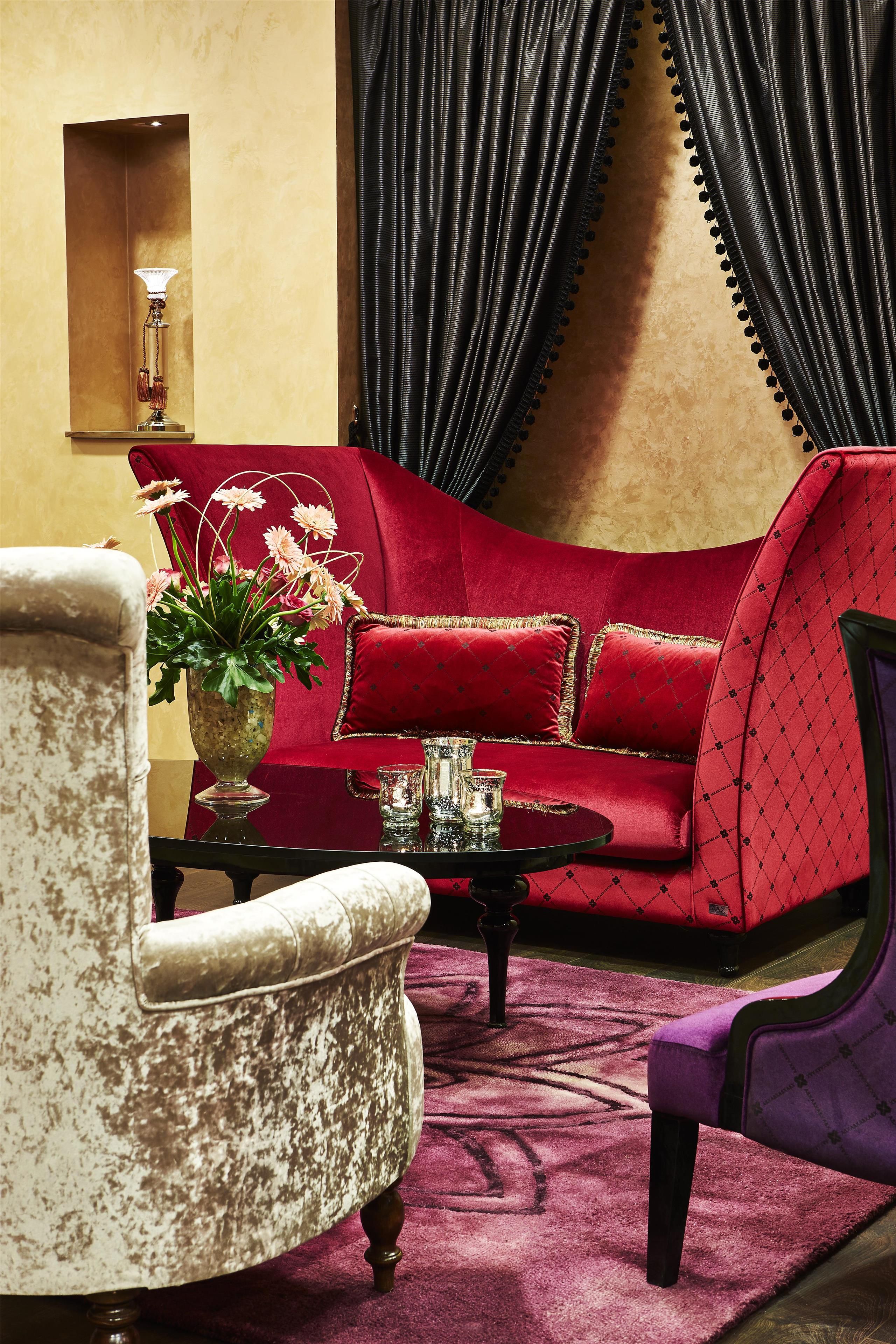 A room with a red sofa, a white armchair, a purple armchair, a small table with a vase of flowers, and a wall-mounted lamp.