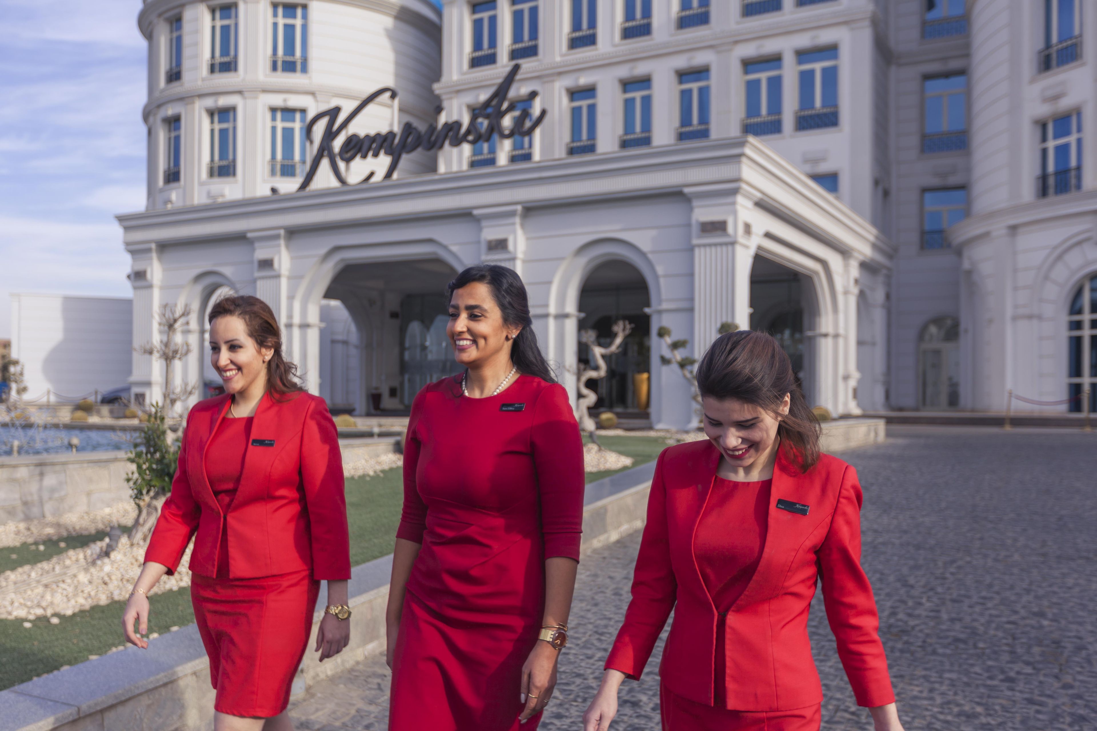 Three individuals in red uniforms walking in front of a building with a sign that reads 'Kempinski'.