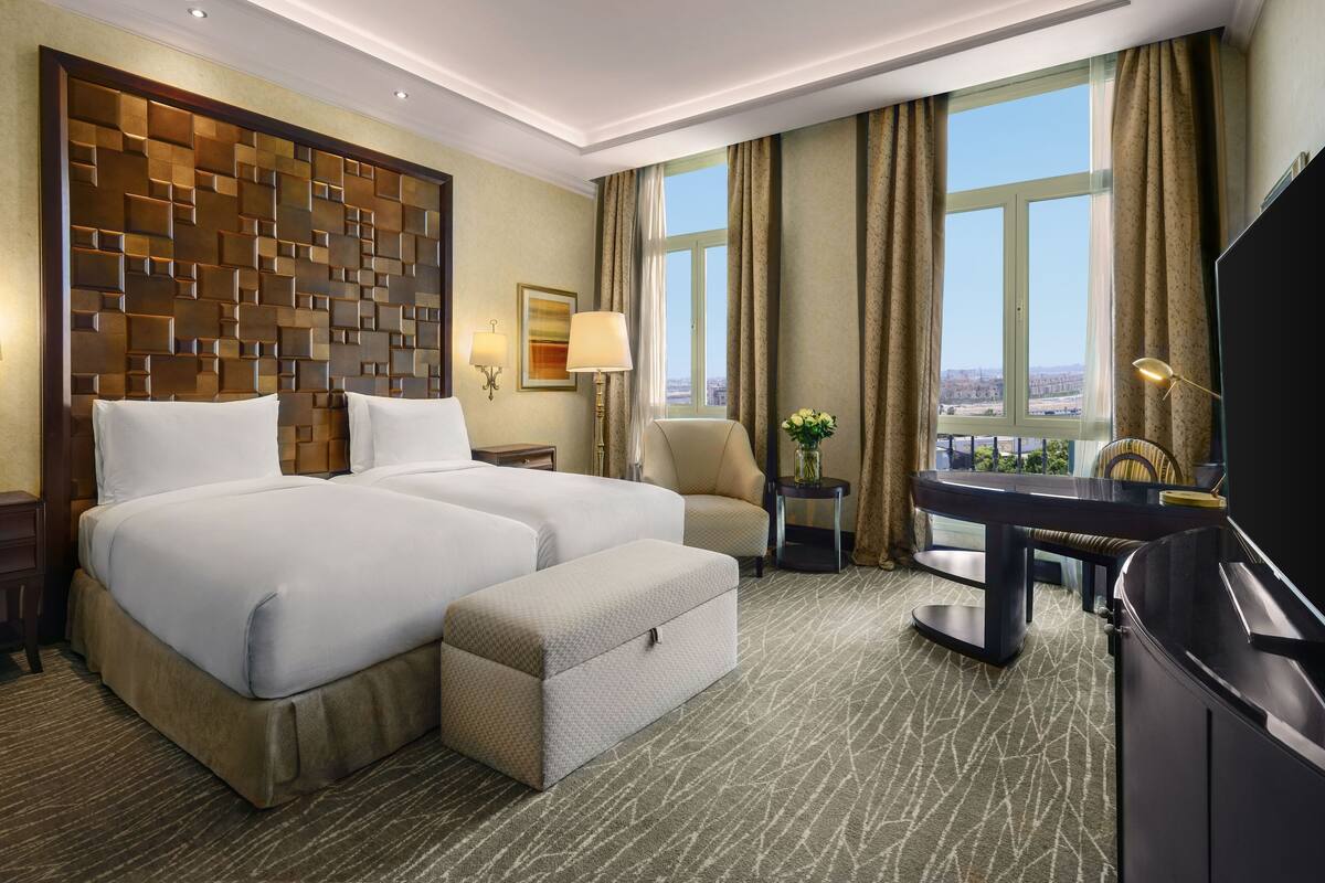 A hotel room with a large bed, a desk, a chair, a TV, and a view of the city.