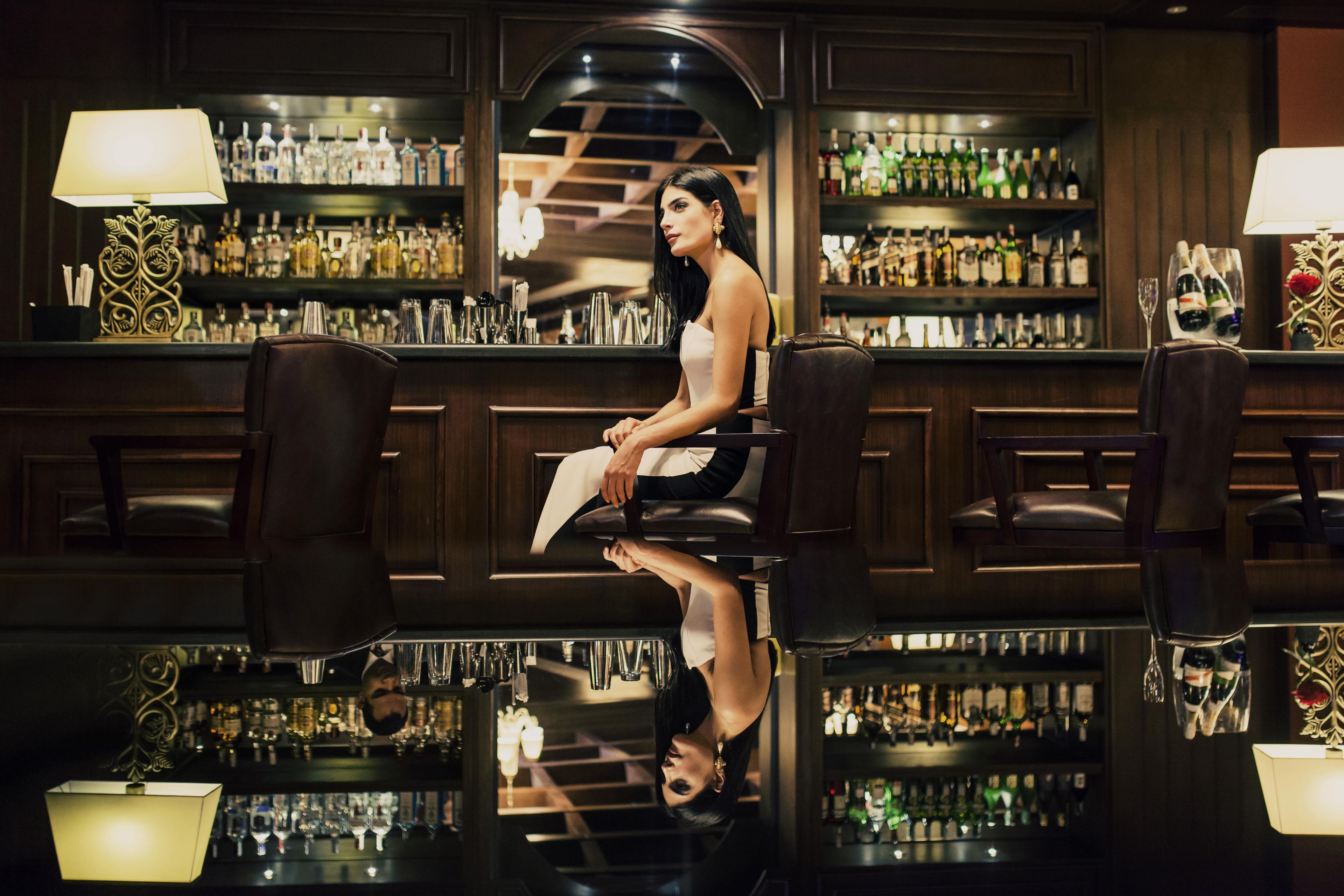 Een bar met een vrouw zittend, omringd door flessen en glazen.