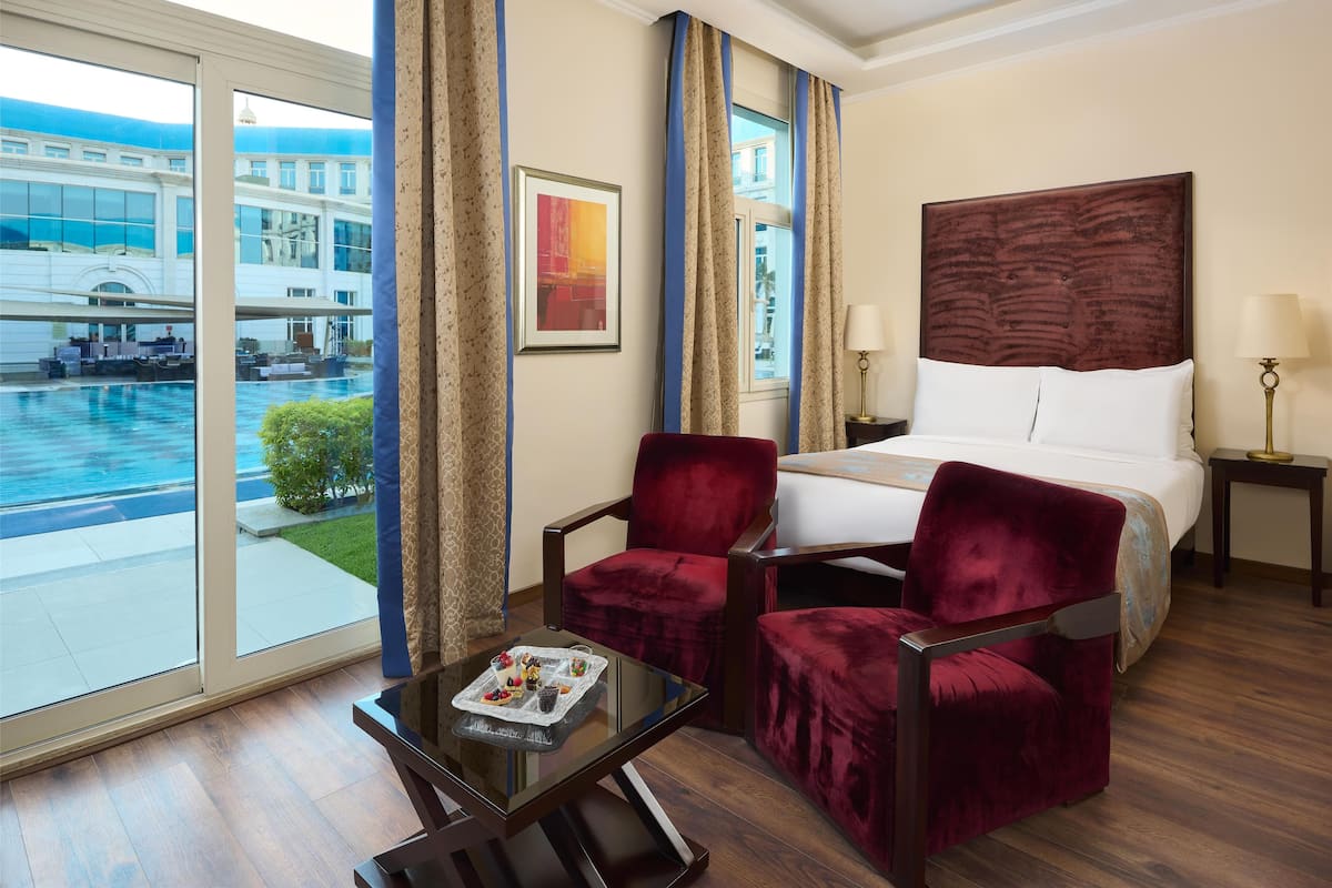 A hotel room with a large window, a bed, two red velvet chairs, a small table with a tray of snacks, and a framed picture on the wall.