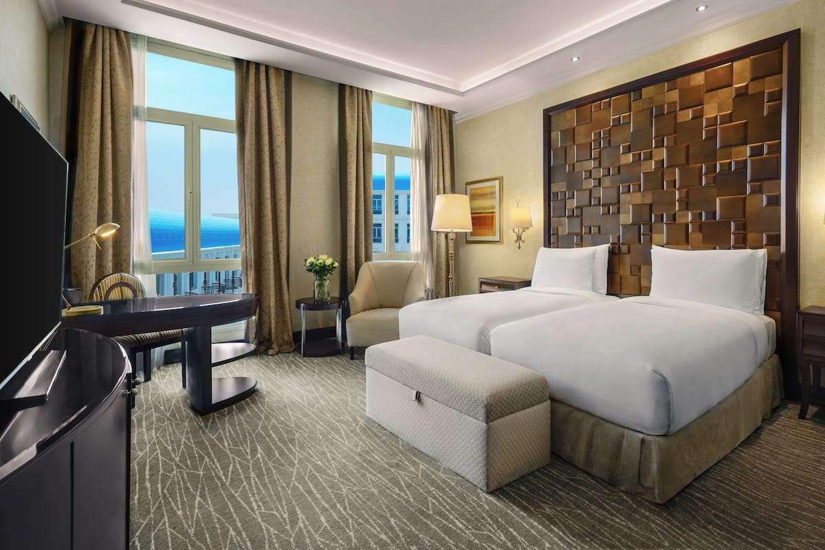 A hotel room with two beds, a desk, a chair, a TV, and a view of the ocean.