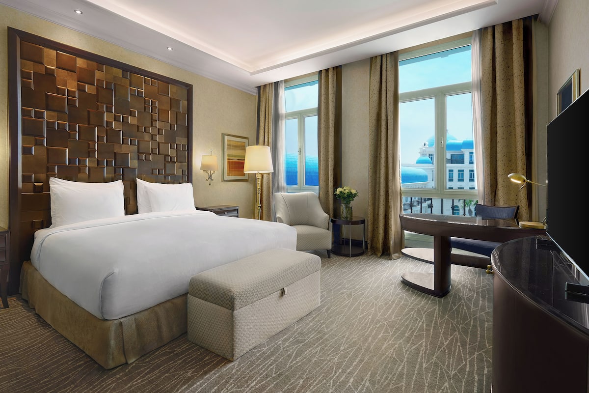 A hotel room with a large bed, a desk, a chair, a TV, and a view of the city.