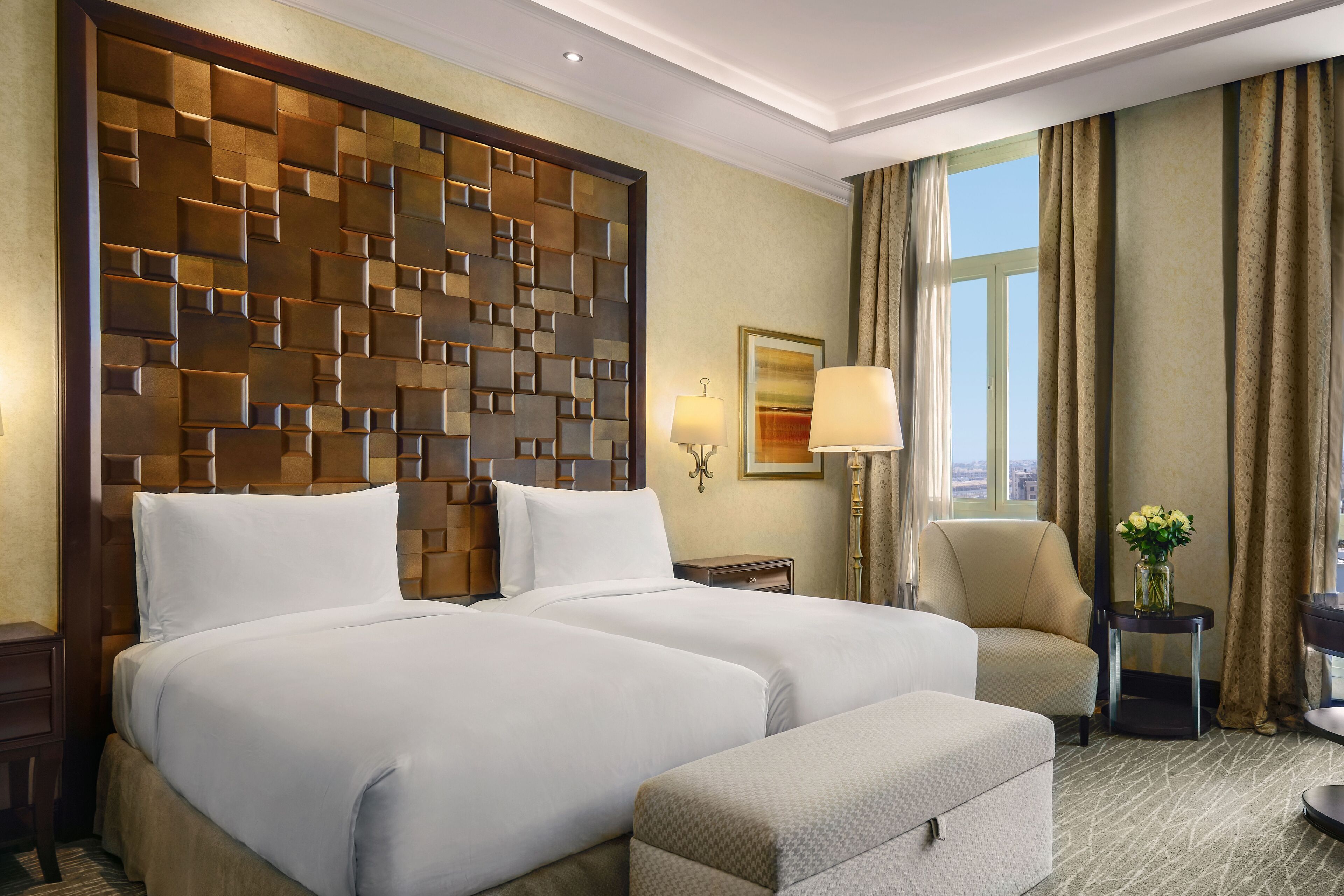 A hotel room with a large bed, a chair, a small table with a vase of flowers, and a window with curtains.