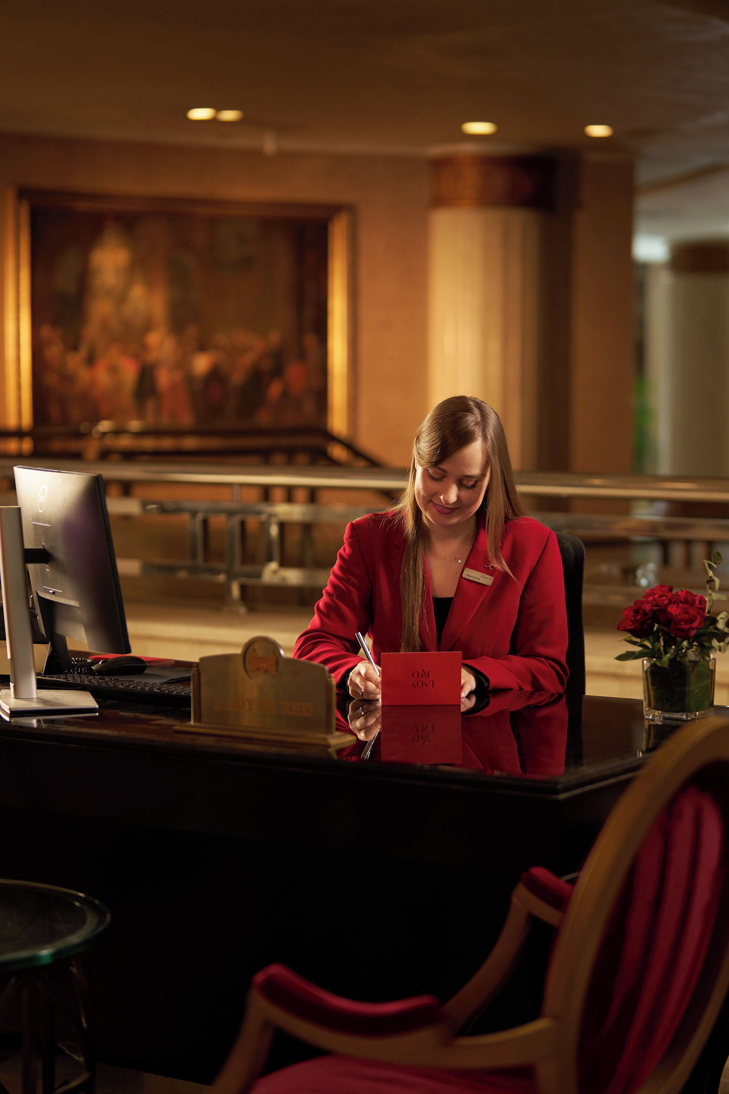A person seated at a desk with a computer, a nameplate, and a red box.