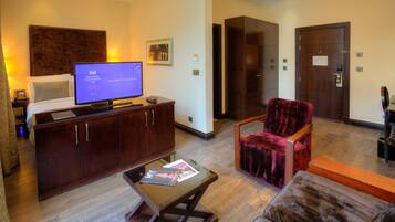 Habitación de hotel con cama, televisor, mueble de madera, una butaca de terciopelo rojo, una mesa de centro de vidrio y suelo de madera.