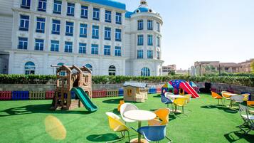 A playground with slides and climbing structures, surrounded by colorful chairs and tables, in front of a large white building with blue accents.