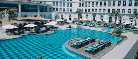A large hotel with a central swimming pool, outdoor seating, and palm trees.
