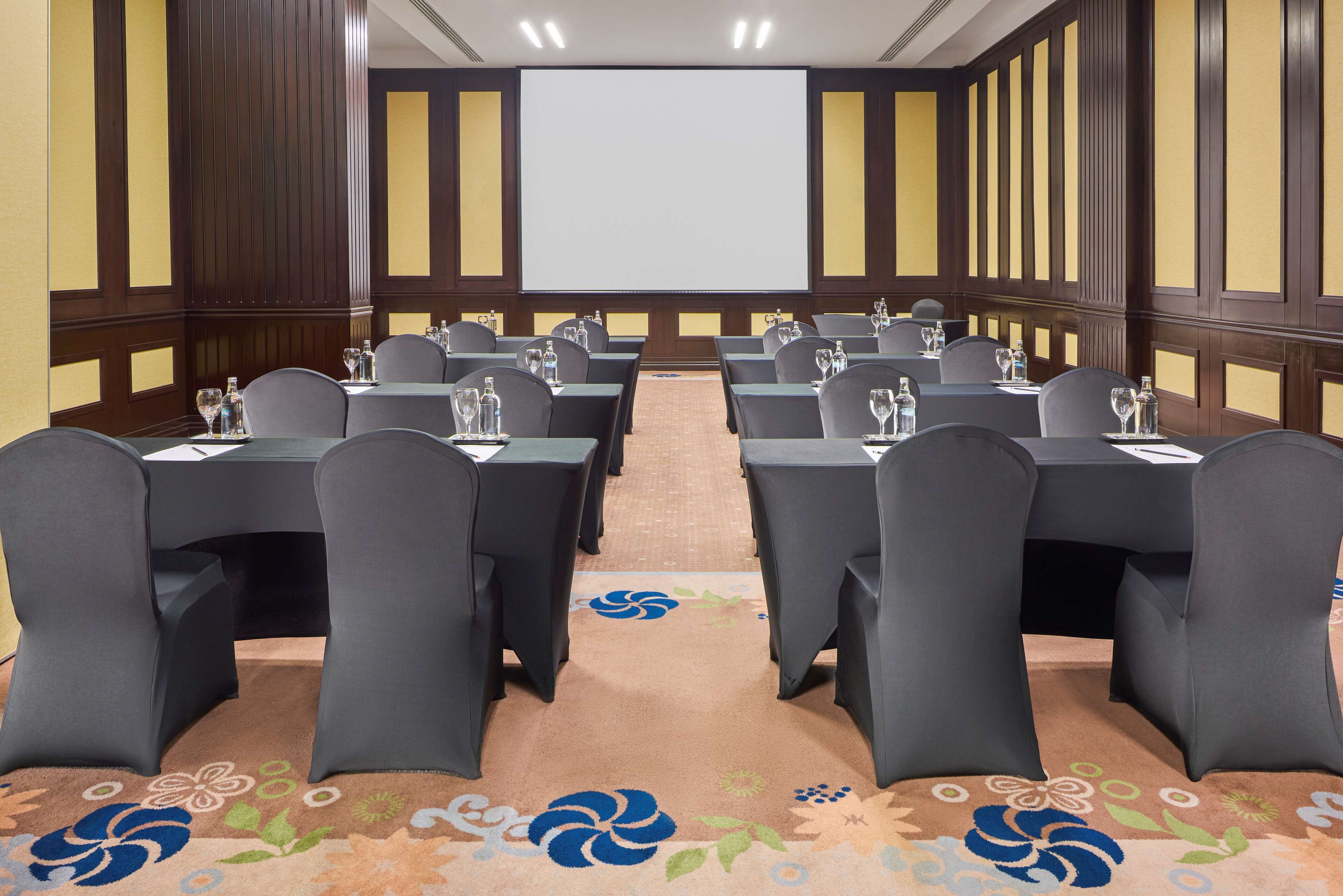 A conference room with rows of tables and chairs, a projector screen, and a carpet with a floral pattern.