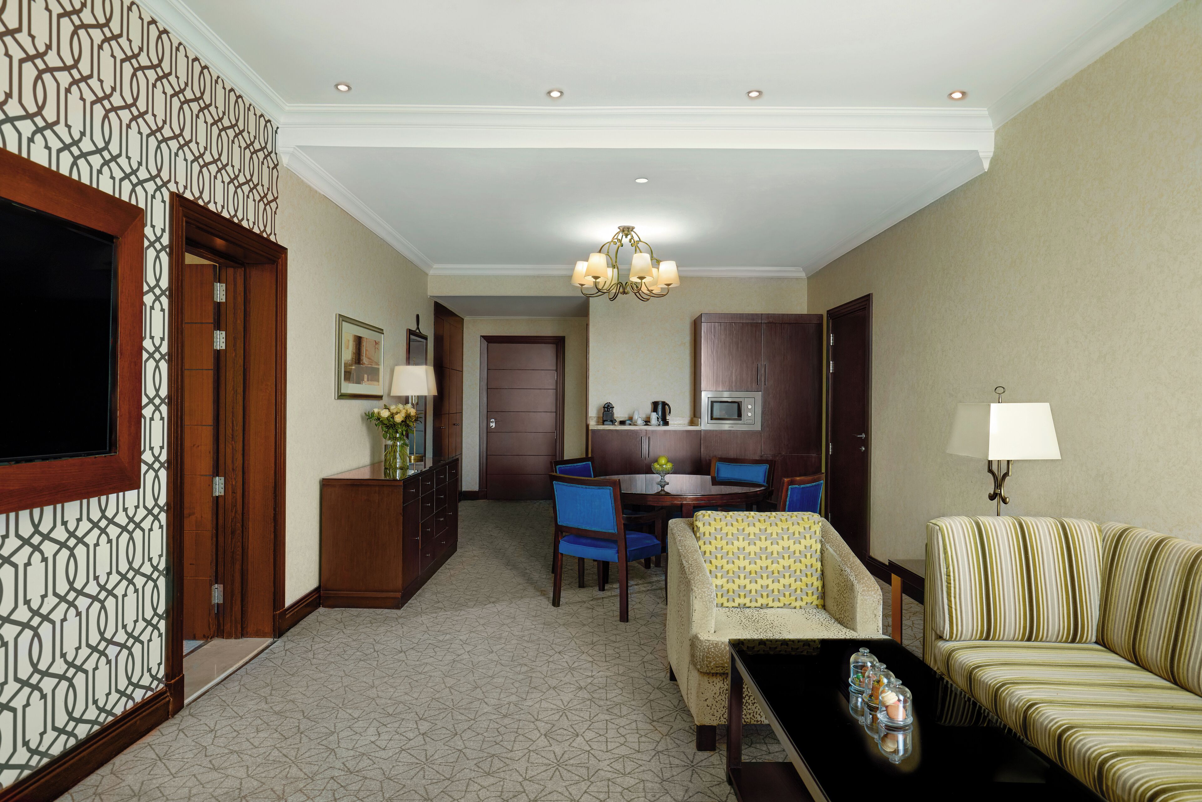 Quarto de hotel com área de jantar, sofá, mesa de centro, televisão e lustre.