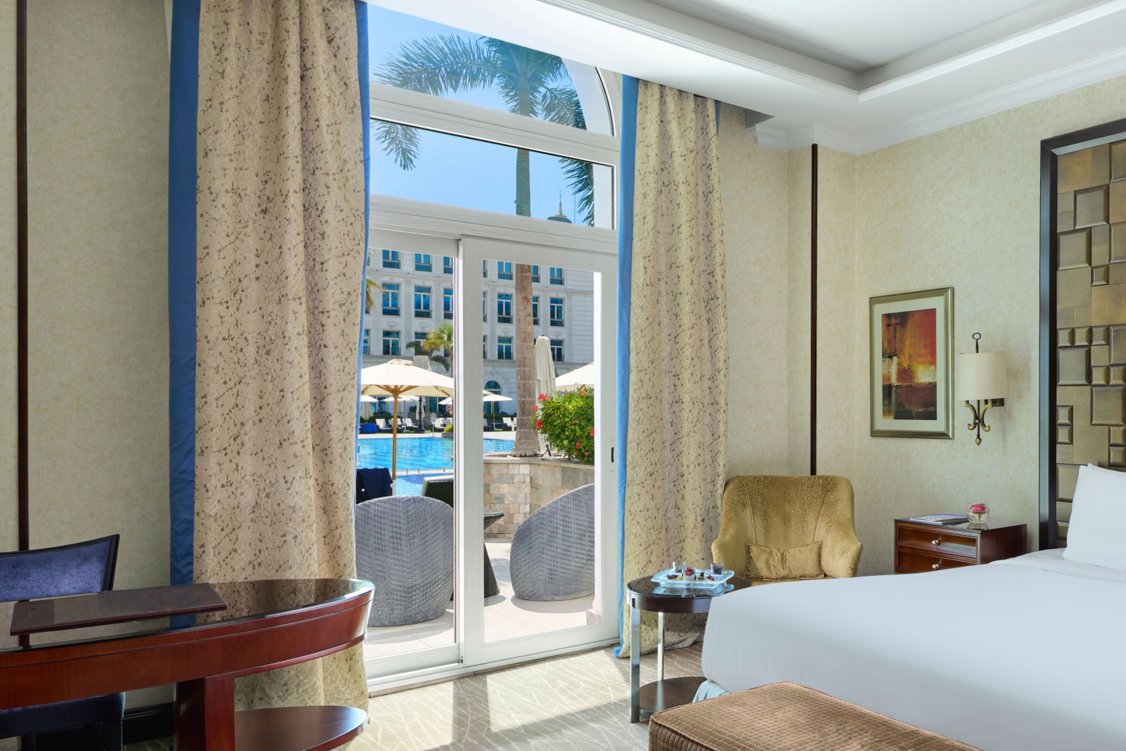 A hotel room with a bed, a desk, a chair, and a view of a pool and buildings outside.