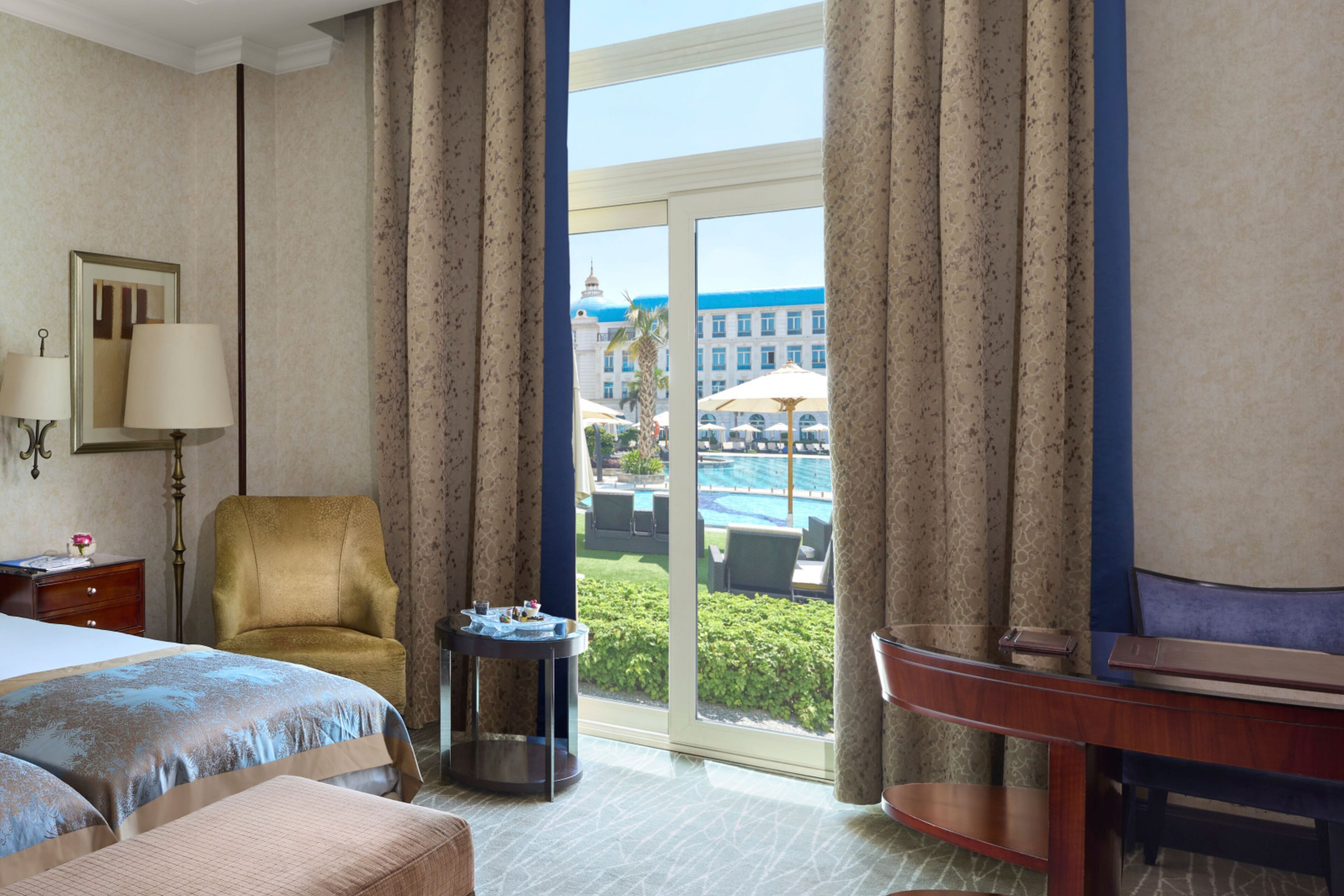 A hotel room with a bed, a chair, a desk, and a view of a pool and building.