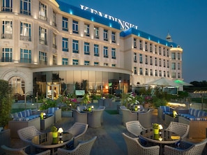 A hotel with a large outdoor seating area featuring wicker furniture and tables, a well-lit entrance, and a prominent building facade with a dome structure. - Kempinski Palace Cairo (New Cairo)