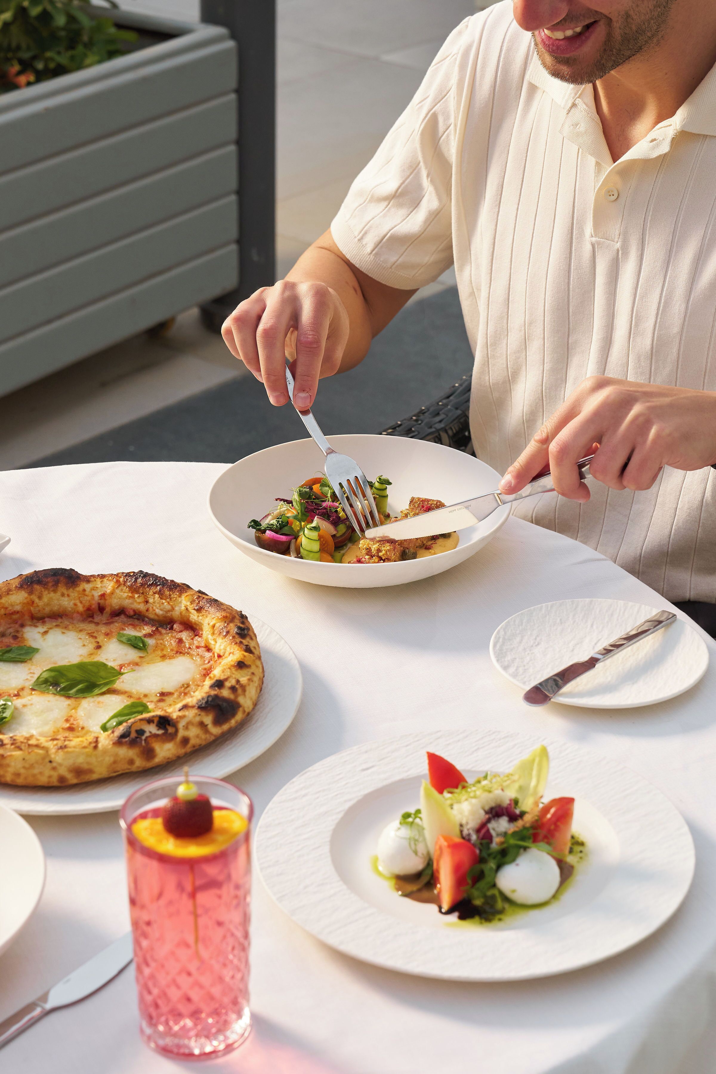 Une personne est assise à une table en plein air et déguste une pizza, une salade et une boisson.