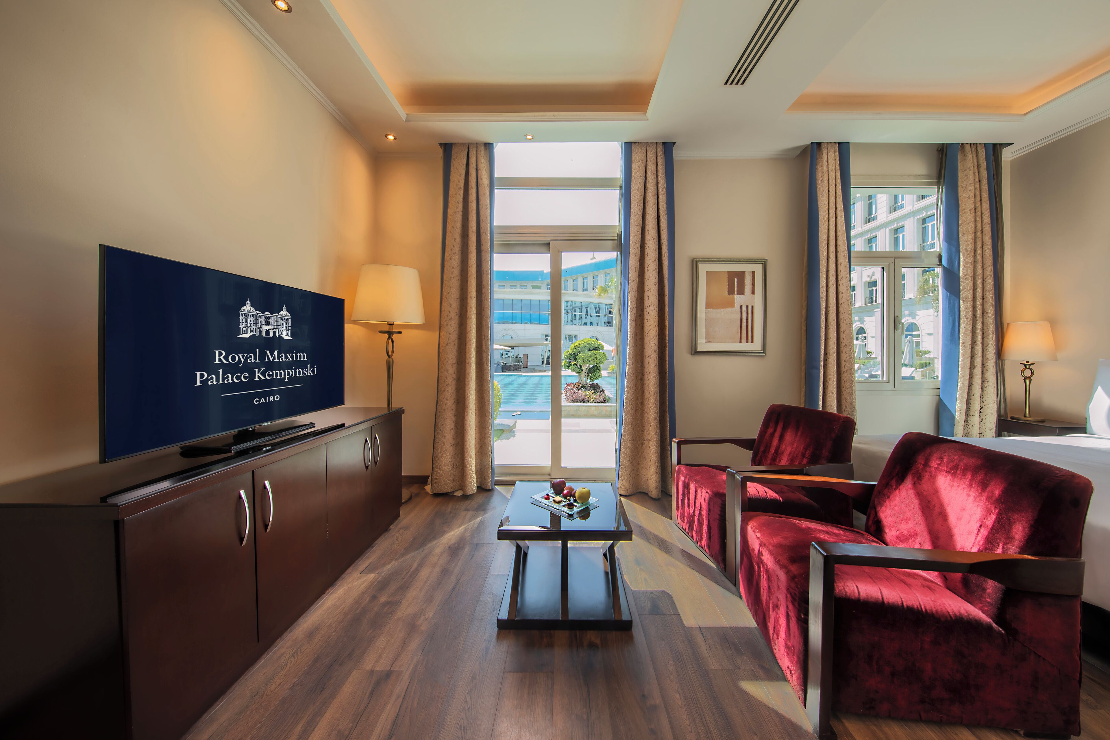 A hotel room with a flat-screen TV displaying the Royal Maxim Palace Kempinski logo, a wooden cabinet, a coffee table, two red velvet armchairs, and a large window with curtains.