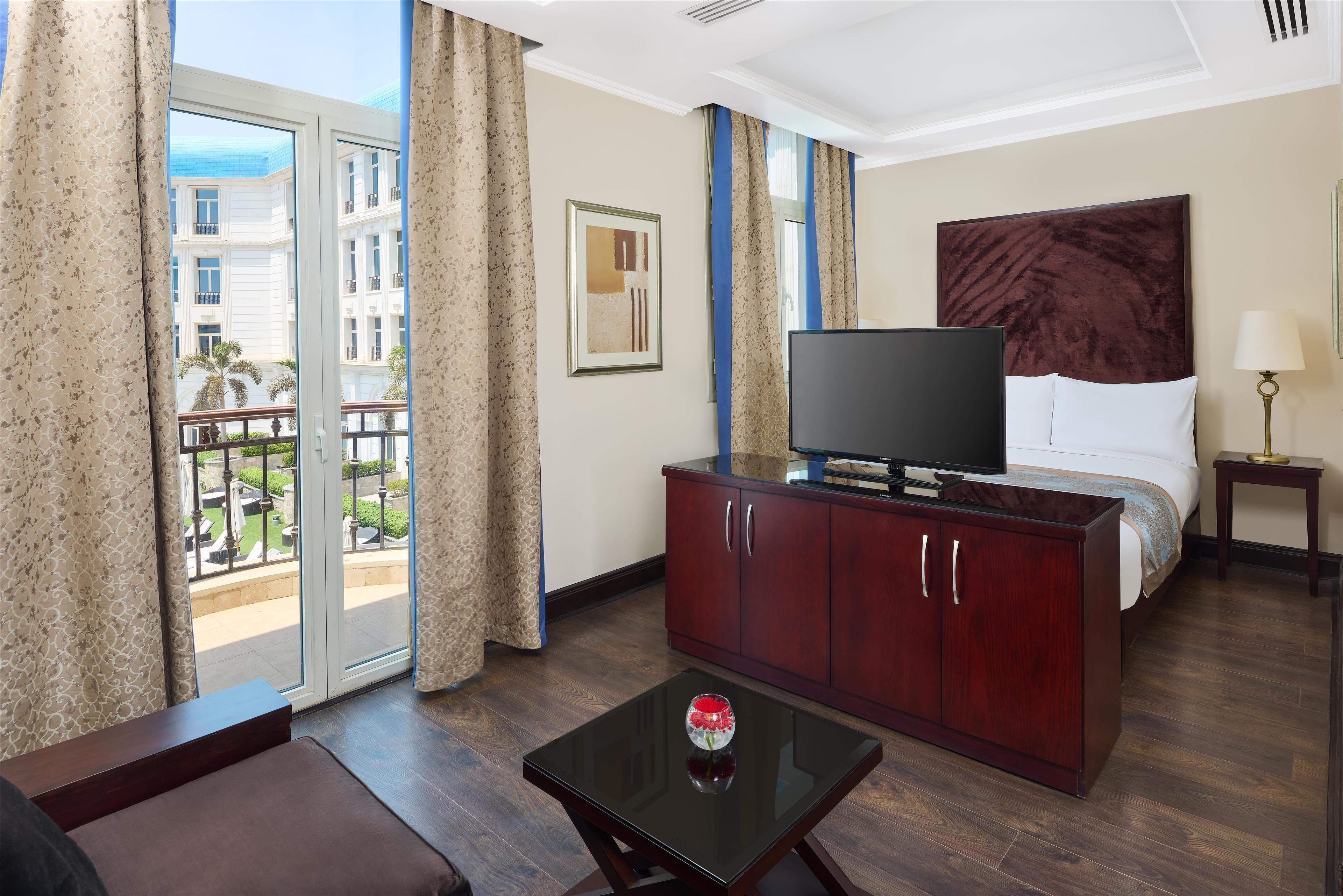 A hotel room with a bed, a TV on a wooden cabinet, a small table with a vase, and a balcony with curtains.