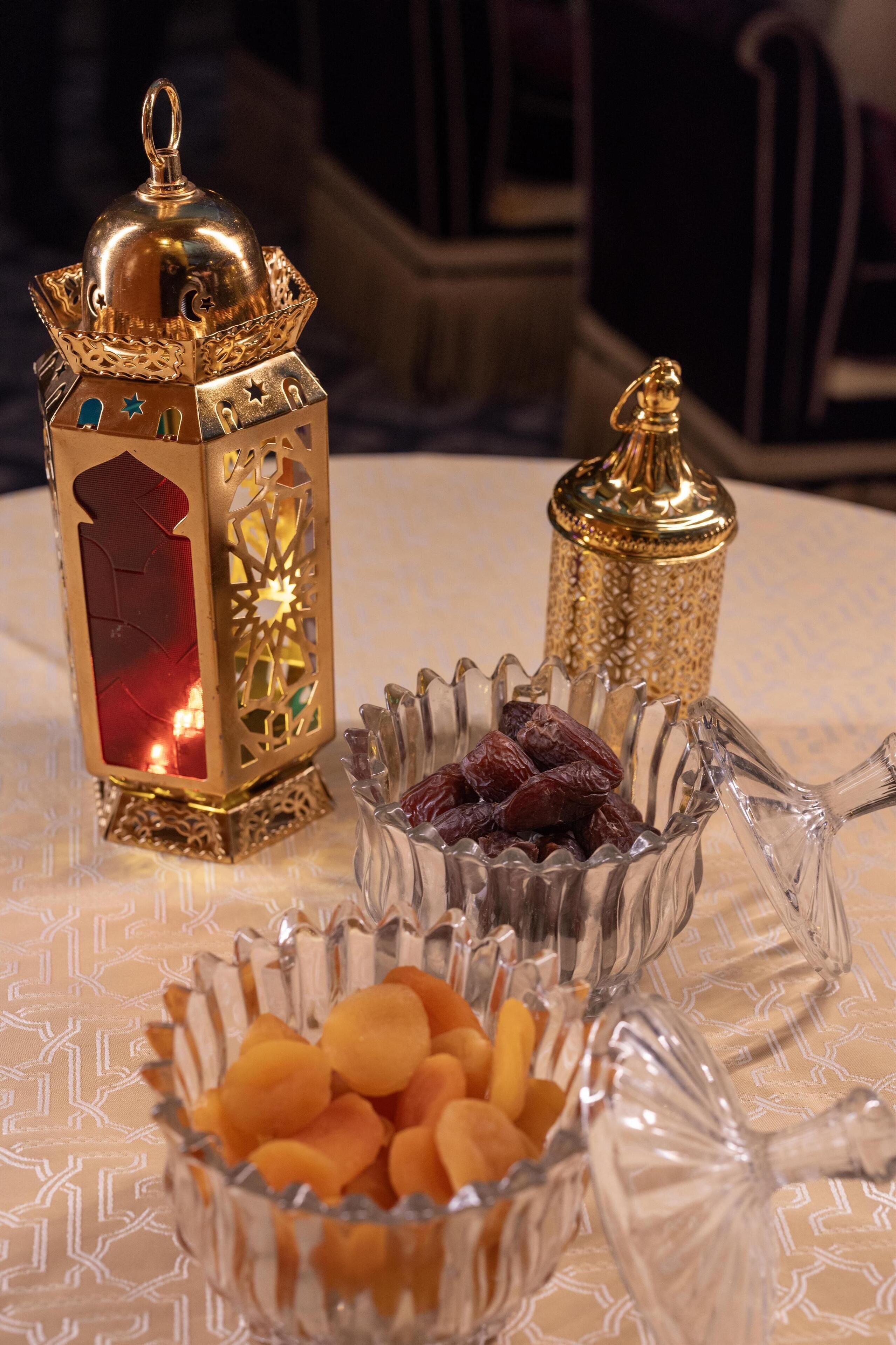 Una mesa con un farol decorativo, un cuenco de vidrio con dátiles y un cuenco de vidrio con frutas secas.