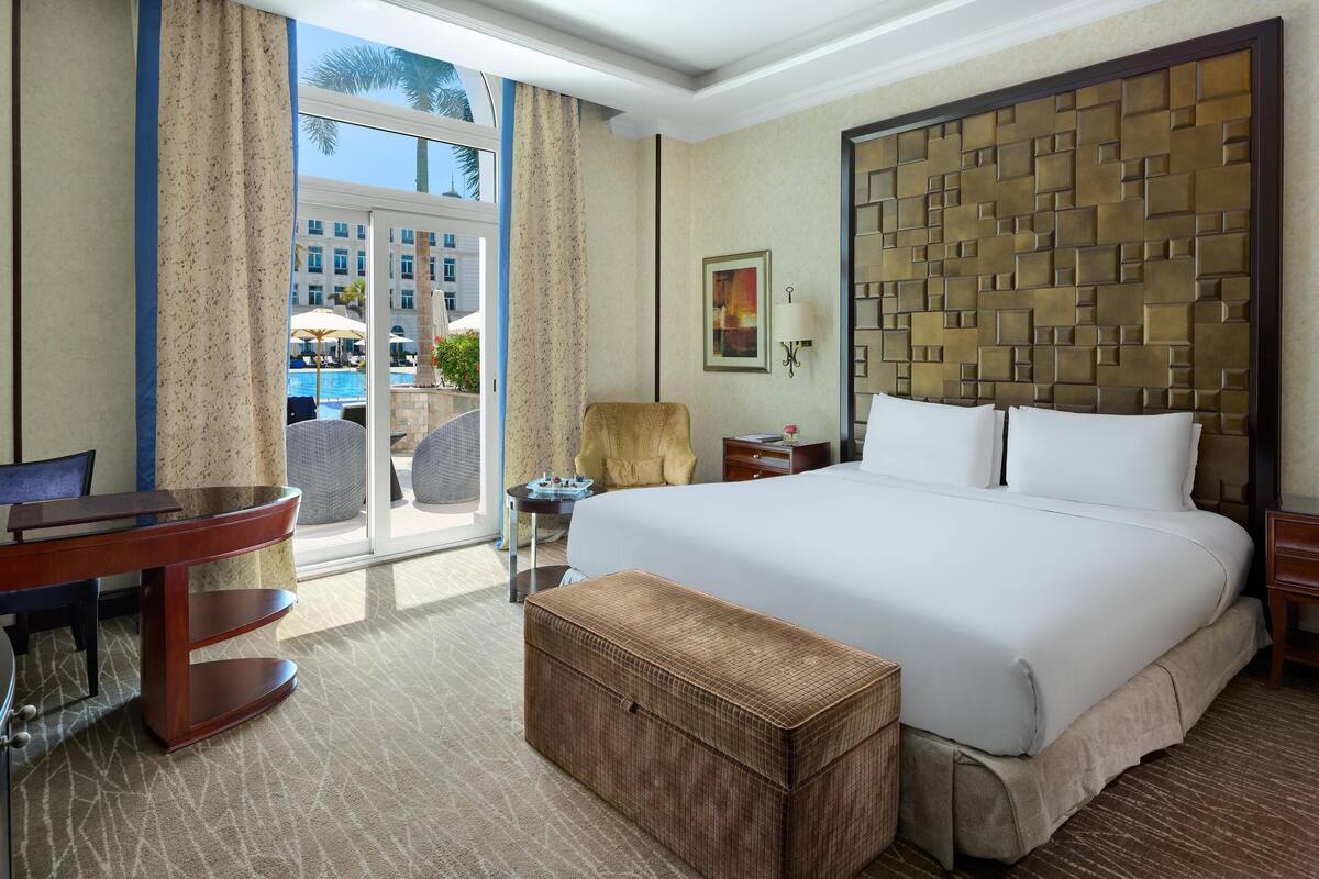 A hotel room with a large bed, a desk, a chair, a small table, a wicker ottoman, and a view of a pool and palm trees through a glass door.
