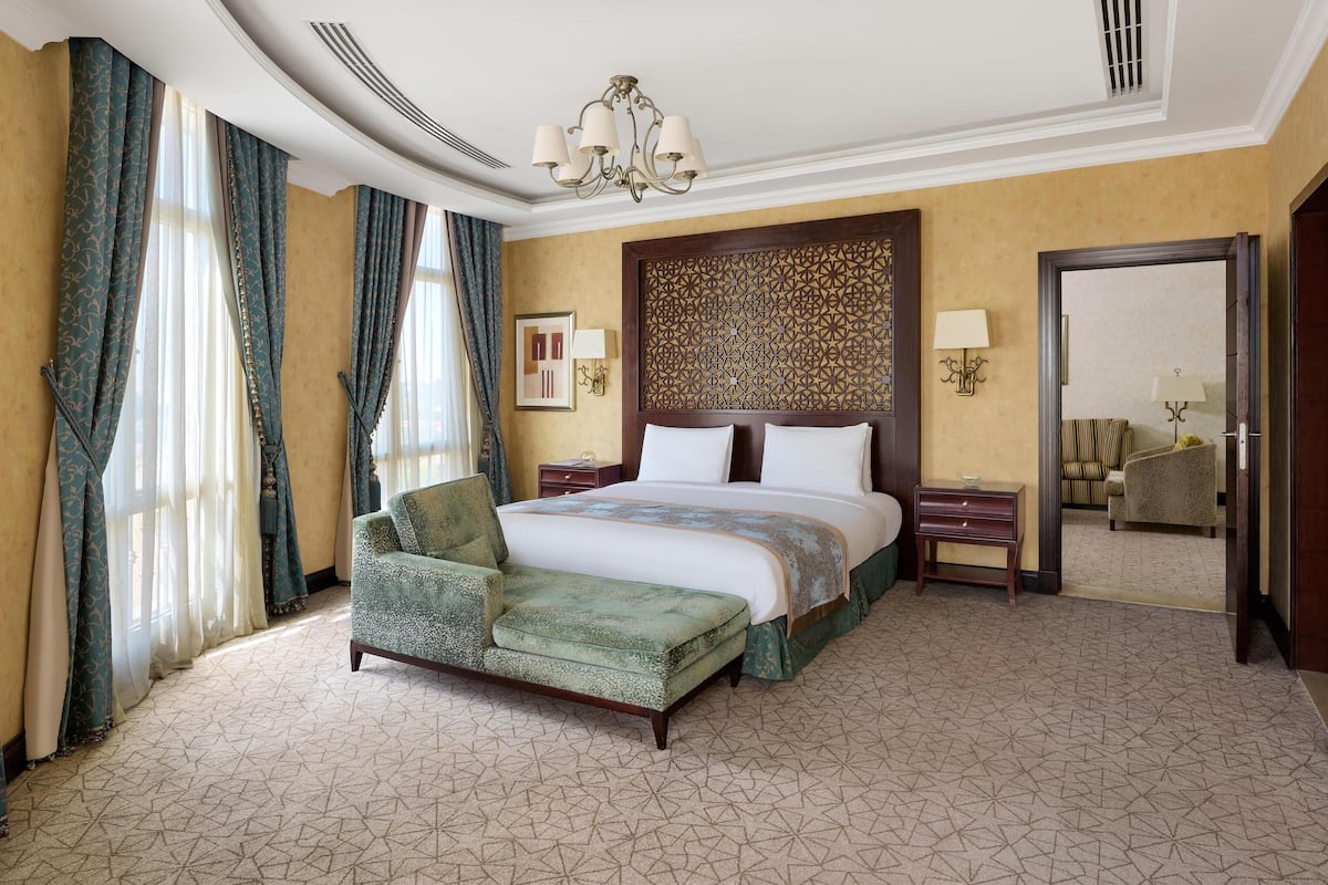 A hotel room with a large bed, a green upholstered armchair, a nightstand, a wall-mounted lamp, and a decorative panel on the wall.