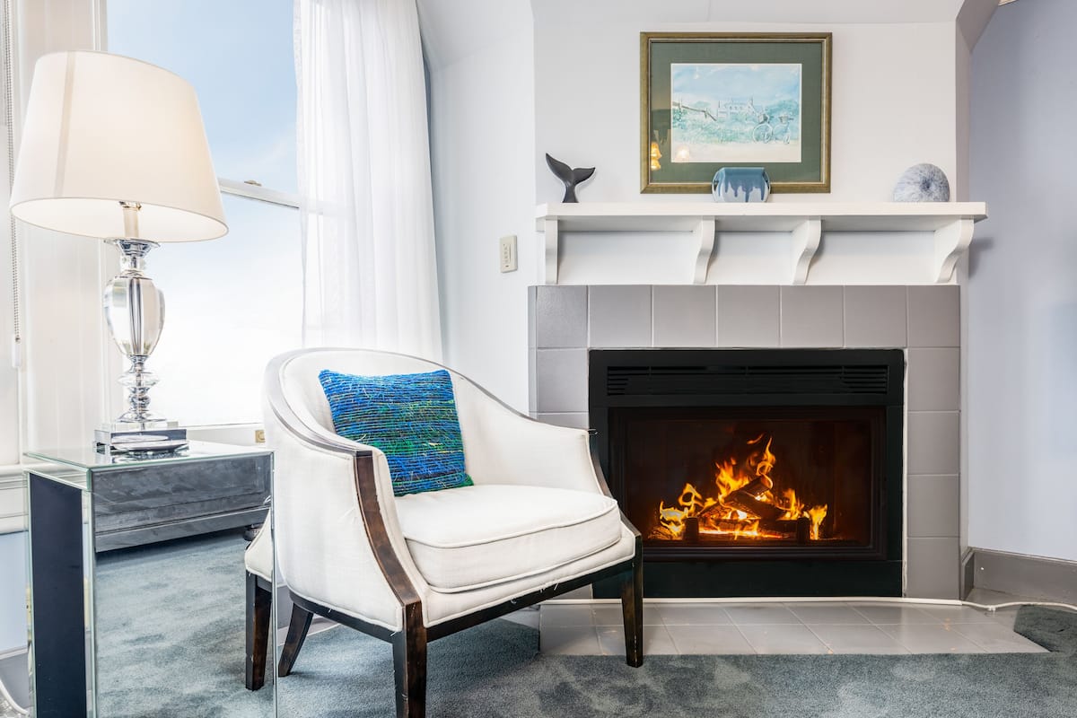 A modern living room with a white armchair, a glass side table, a fireplace, and a framed picture above the mantel.