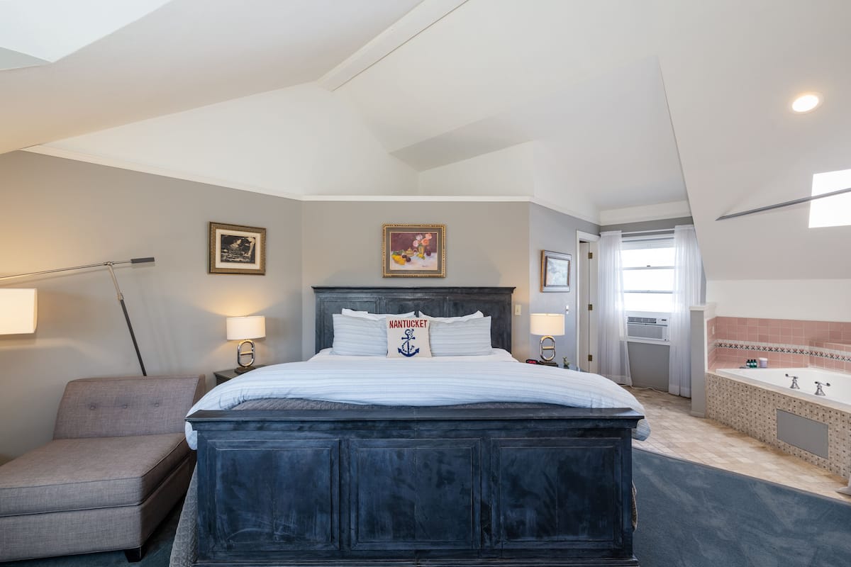 A hotel room with a large bed, a bedside table with a lamp, a chair, a bathroom with a bathtub, and a window with curtains.