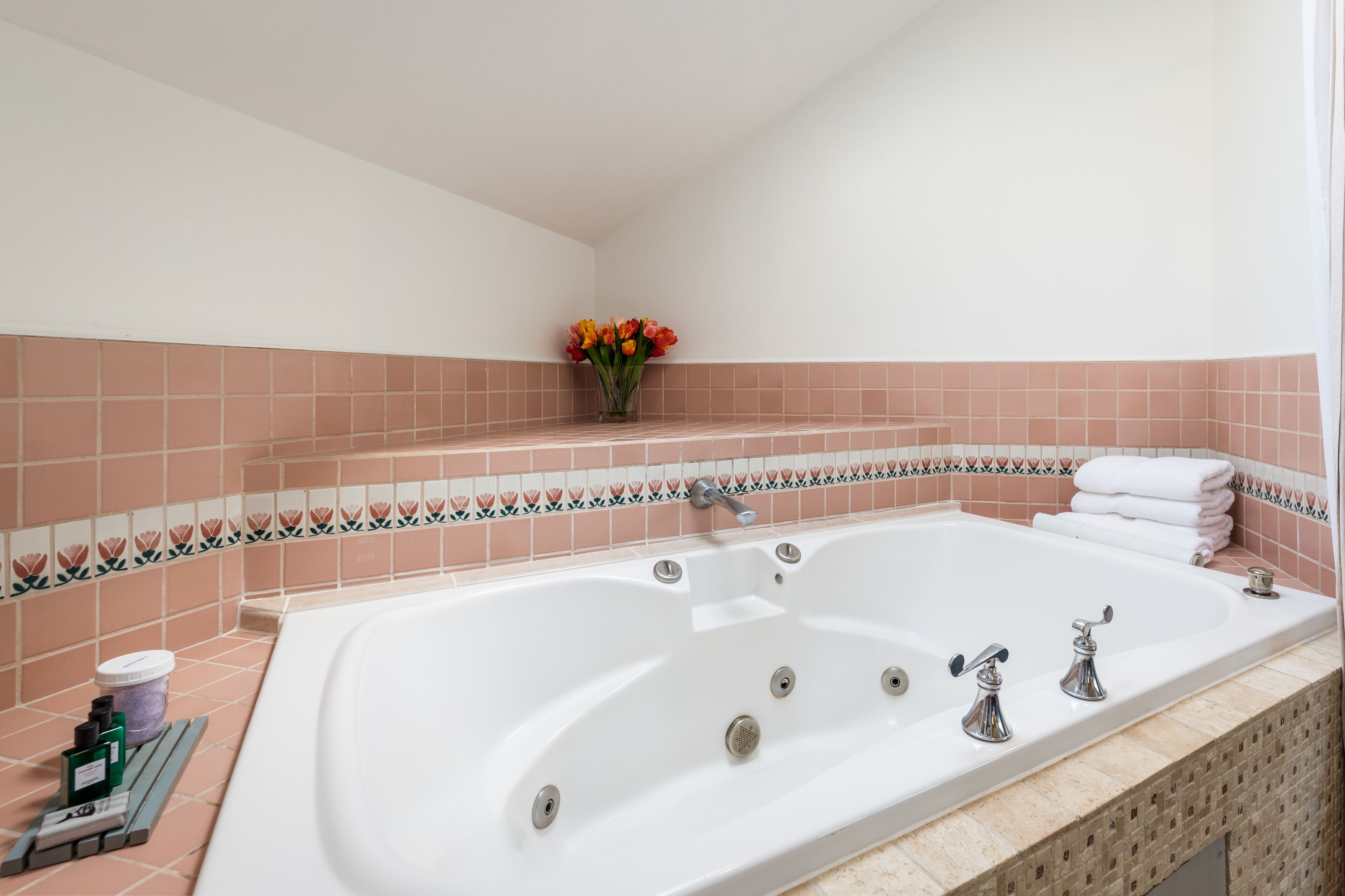 A bathroom with a large bathtub, pink tiled walls, a small vase with flowers, and neatly stacked white towels.