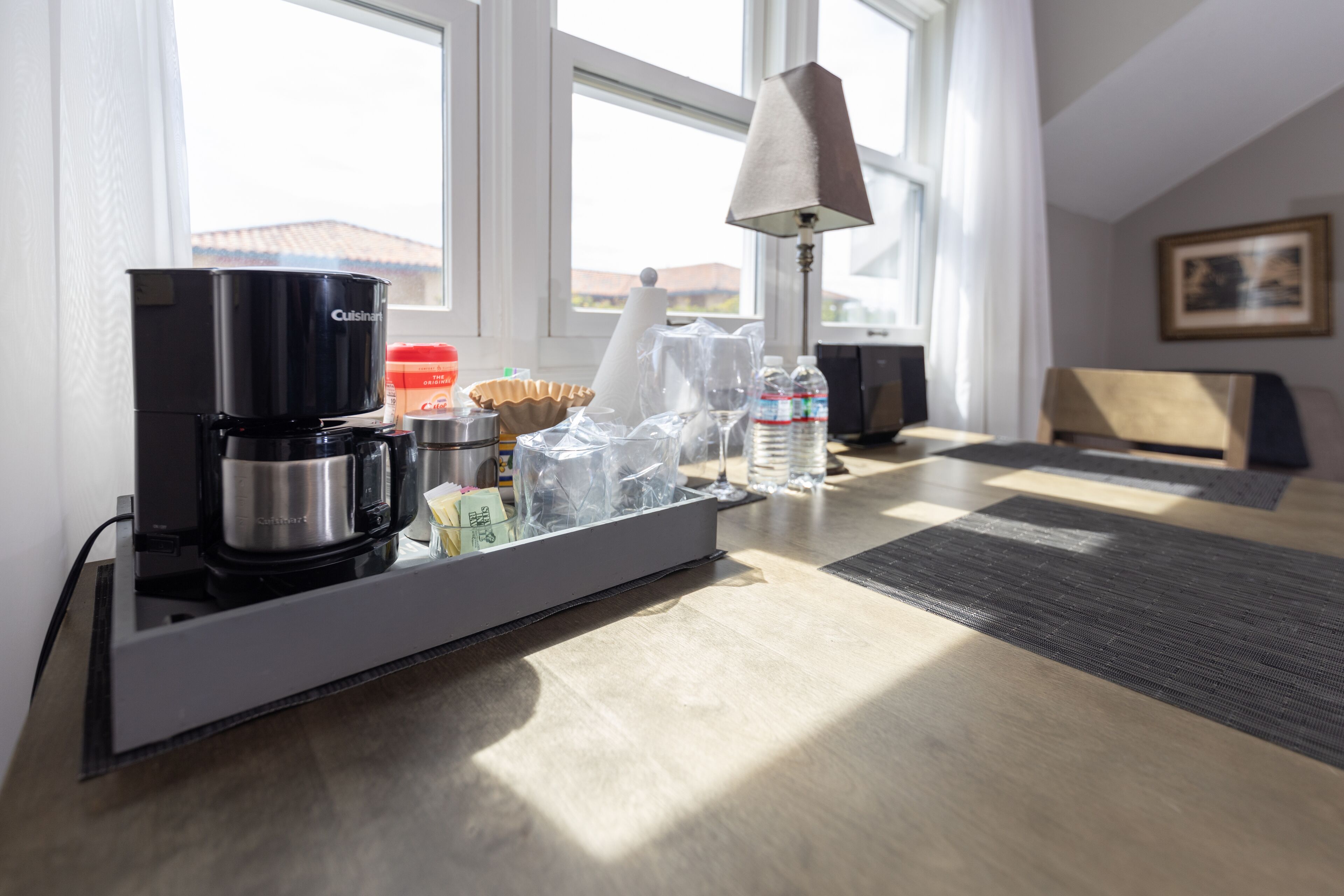 A hotel room with a kitchenette area, a coffee maker, a lamp, and a view of the outside.