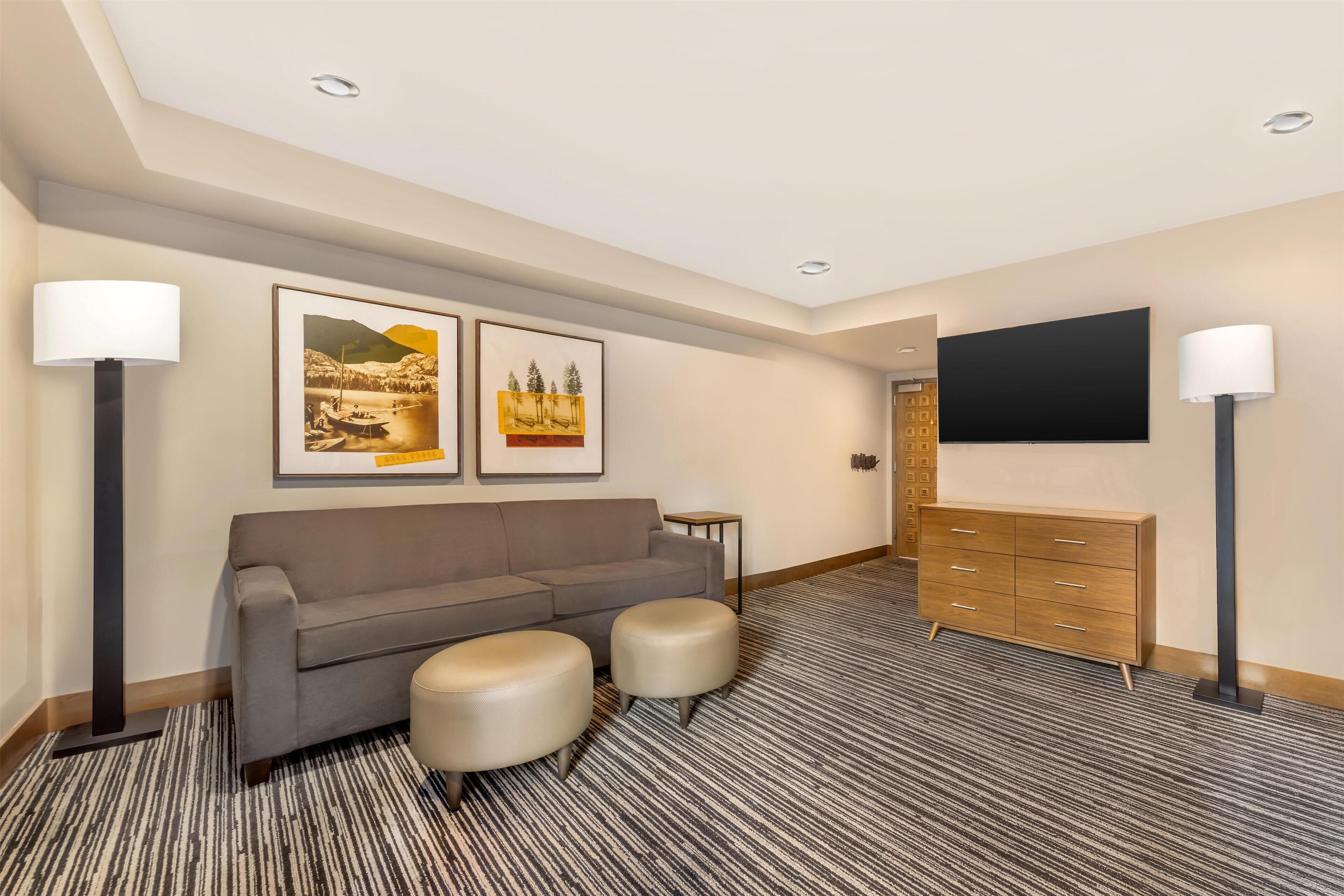 Ein Hotelzimmer mit grauem Sofa, zwei Hocker, einer Holzkommode und einem an der Wand befestigten Flachbildfernseher.