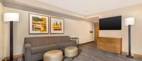 A hotel room with a grey sofa, two ottomans, a wooden dresser, and a flat-screen TV mounted on the wall.