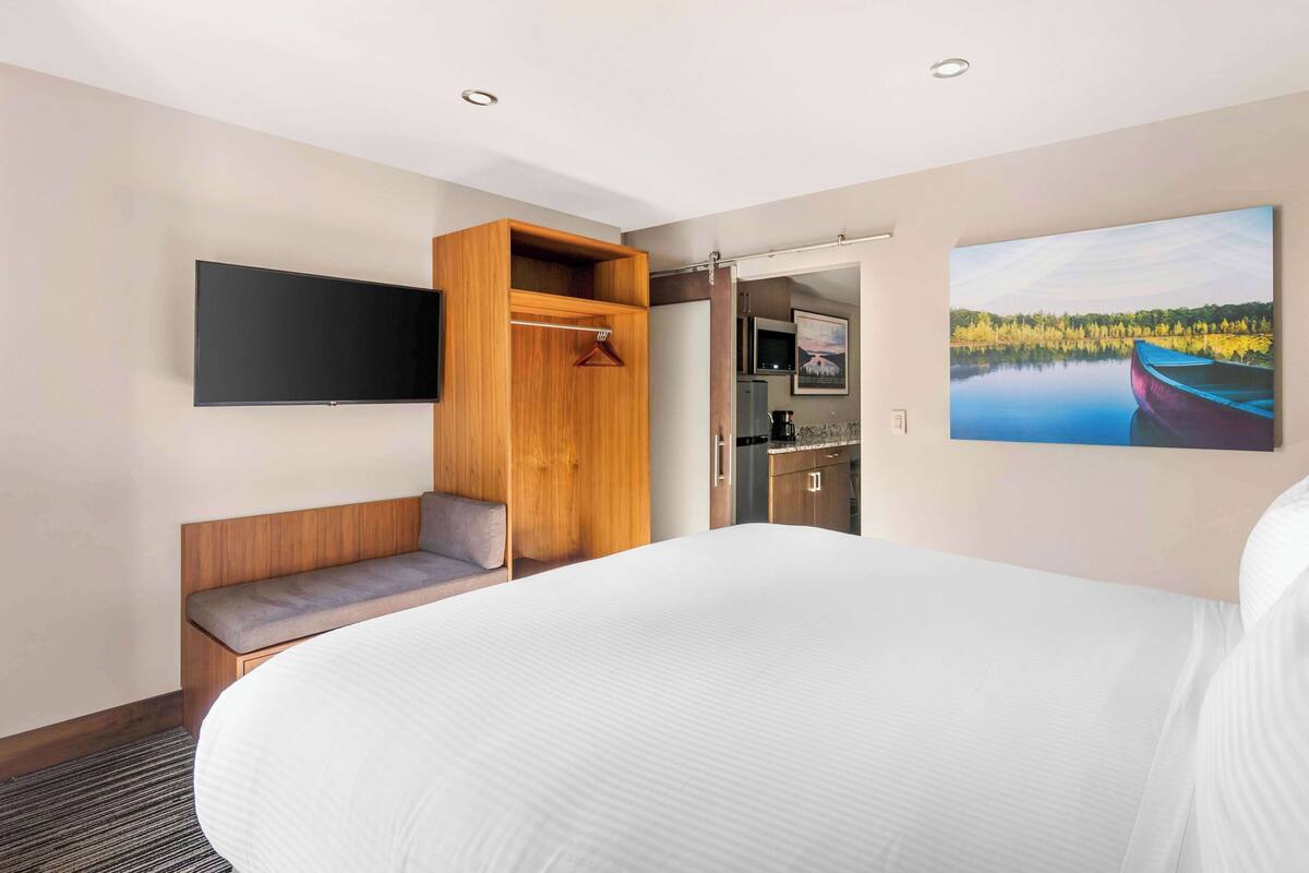 Una habitación de hotel con una cama grande, un banco de madera, un televisor montado en la pared, una kitchenette y un cuadro de un lago.