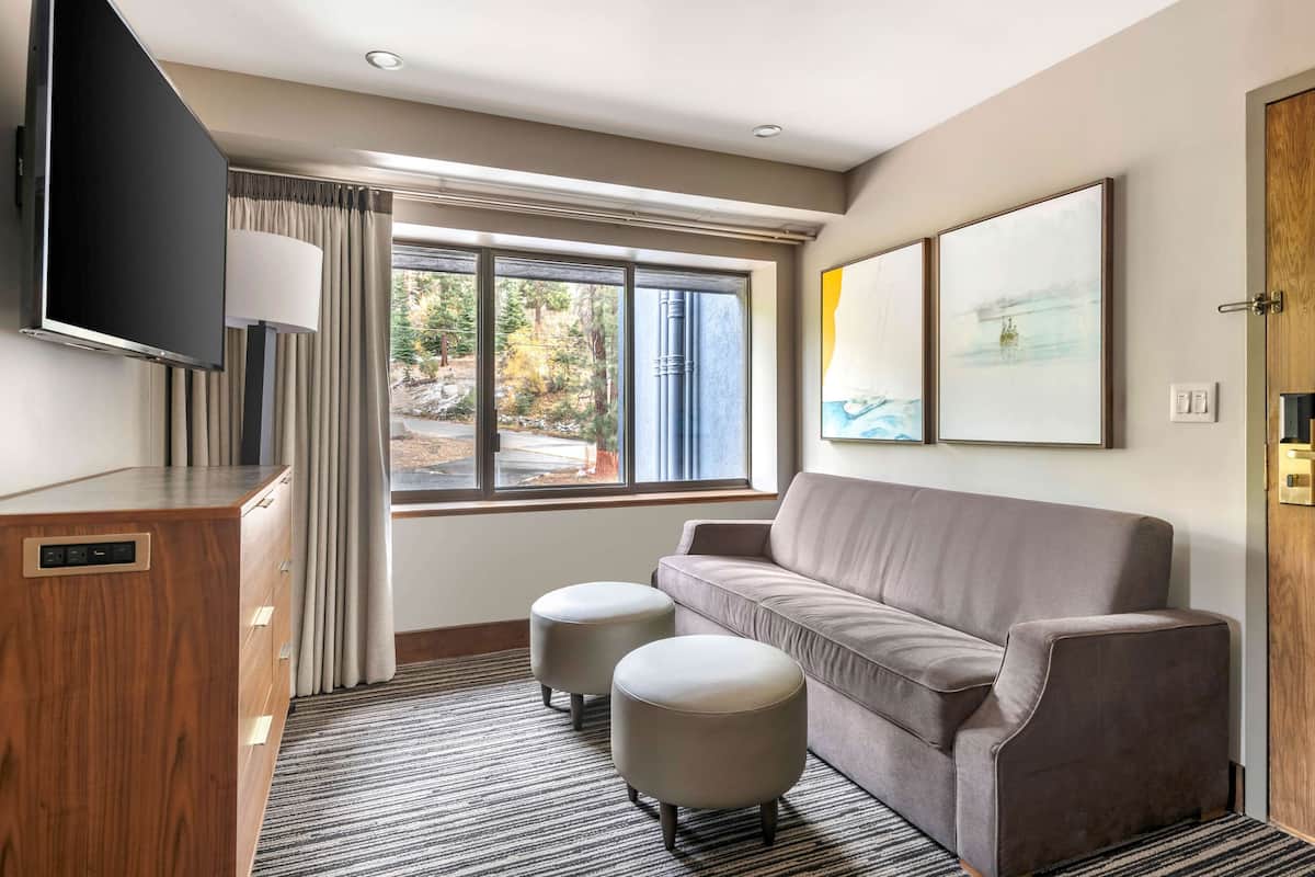 Una habitación de hotel moderna con sofá, otomanes, televisor y ventana con cortinas.