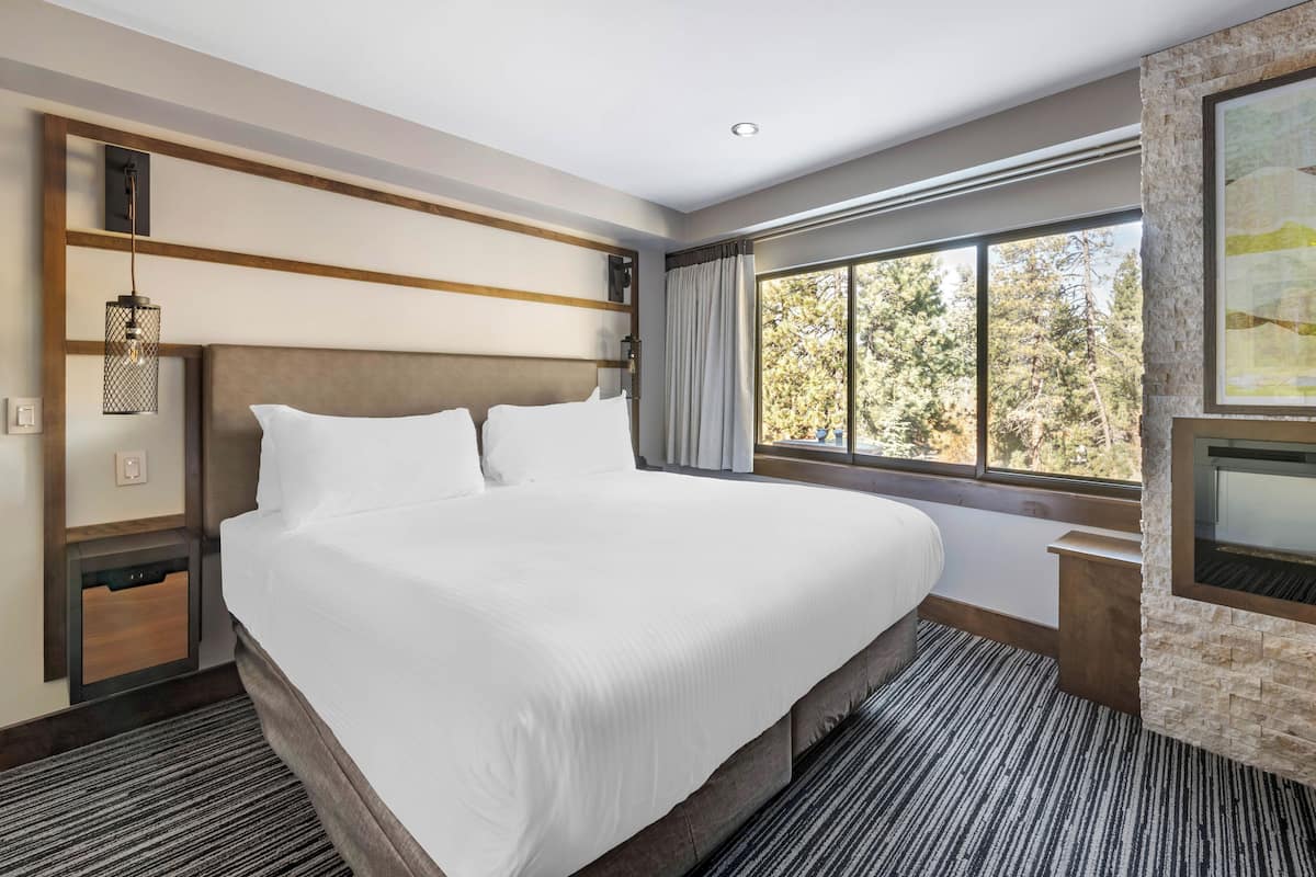 Una cama grande con ropa blanca, cabecera de madera, mesita de noche con lámpara, pared de piedra decorativa y una ventana amplia con vistas a árboles.