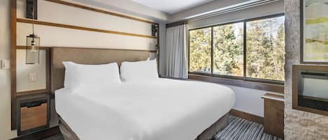 A large bed with white linens, a wooden headboard, a nightstand with a lamp, a stone accent wall, and a large window with a view of trees.