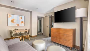 A hotel room with a sofa, ottoman, wooden dresser, flat-screen TV, and a painting on the wall.