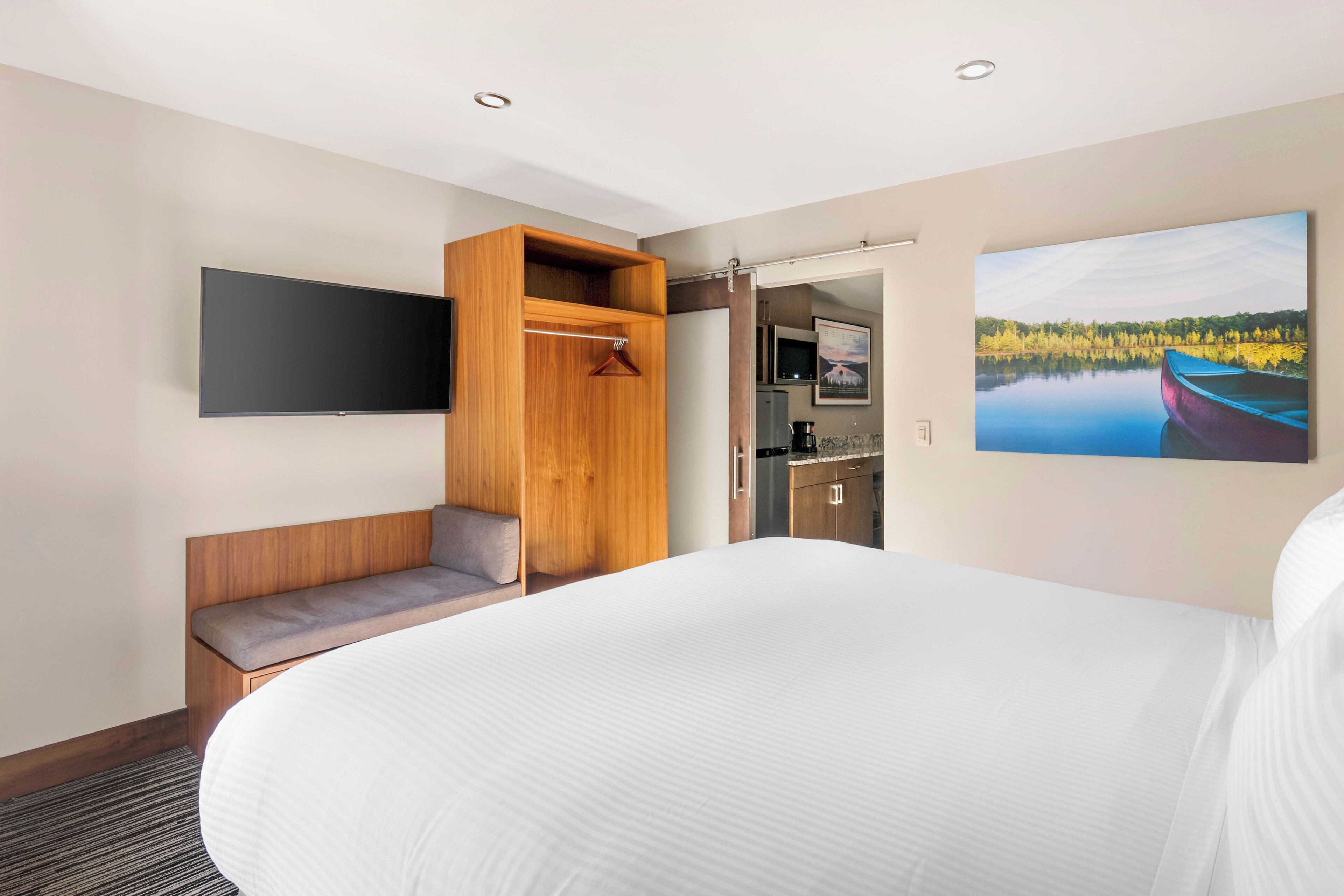 A hotel room with a large bed, a wooden bench, a TV mounted on the wall, a kitchenette, and a painting of a lake.