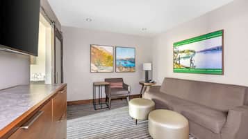 A modern hotel room with a flat-screen TV, a sofa, two ottomans, a small table, and two framed landscape paintings.