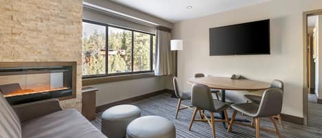 Habitación de hotel con pared de piedra, chimenea, ventanal grande, televisor de pantalla plana, mesa redonda con sillas y sofá.