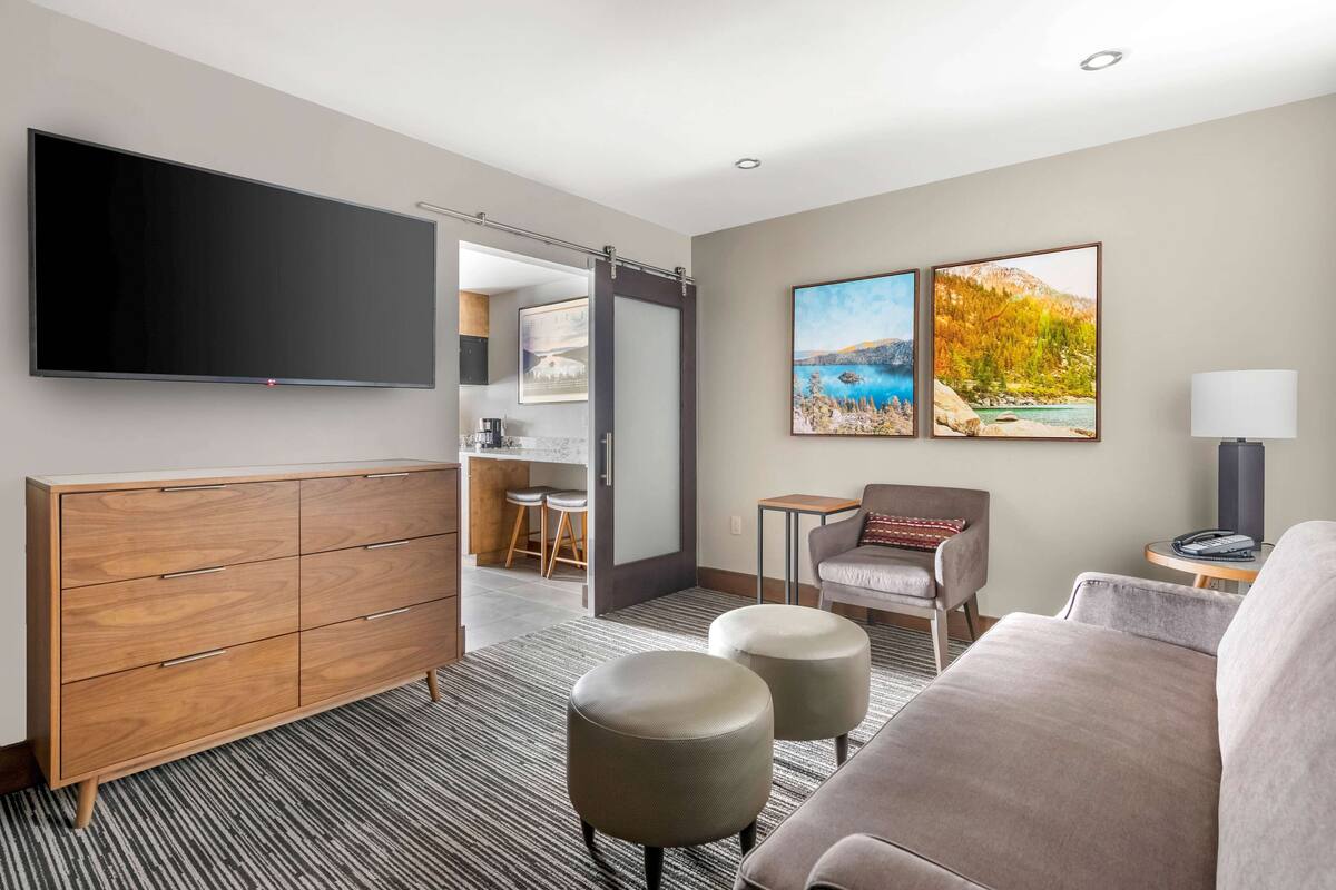 Una habitación de hotel moderna con una televisión de pantalla plana, un mueble de madera, un sofá, dos otomanas, una mesita con una lámpara y dos cuadros enmarcados con paisajes.