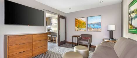A hotel room with a flat-screen TV, a wooden cabinet, a sofa, two ottomans, a chair, and artwork on the walls.
